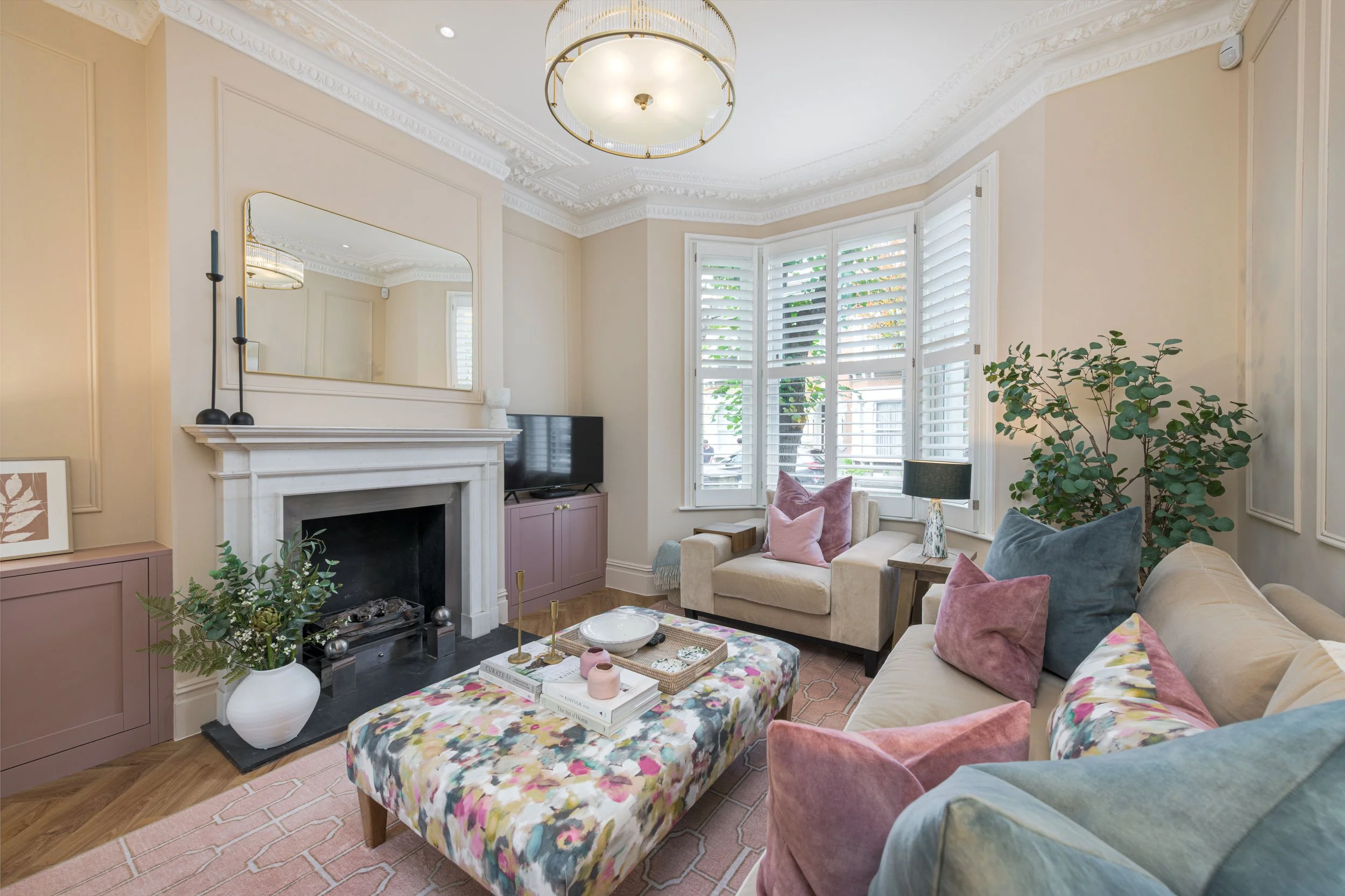 A brightly lit living room with a white fireplace and ornate crown molding ceiling. There is a large window with white shutters, a beige sofa with pink and blue pillows, a pastel pink armchair, and a floral upholstered ottoman. Decor includes a large