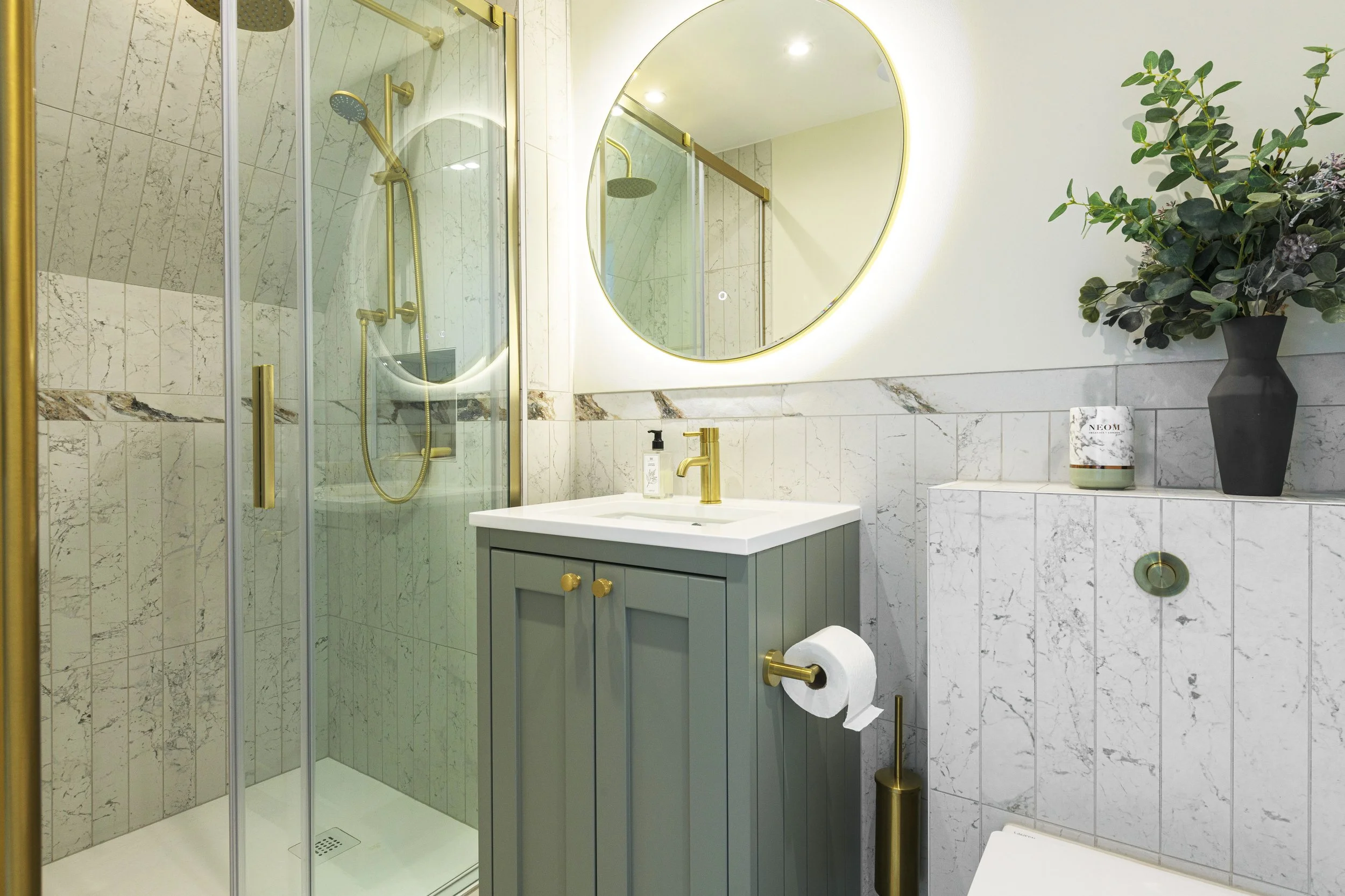 Modern bathroom with marble tiles, a glass shower with brass fixtures, a circular mirror with backlighting, a gray vanity with brass handles, a small potted plant, and a toilet paper holder with a roll of toilet paper.