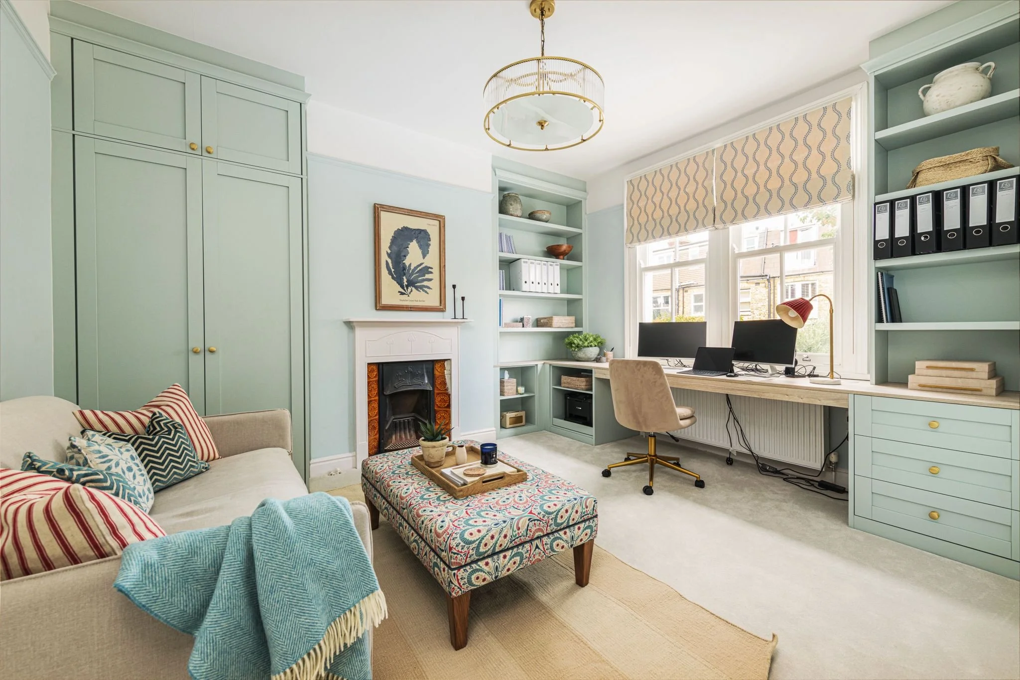 A cozy living room with a light green built-in bookshelf and cabinets, a cream-colored sofa with colorful striped and patterned pillows, a patterned ottoman, and a wall with a fireplace and artwork. There is a beige armchair, a wooden desk with two computer monitors, a red desk lamp, and shelves with decorative items and binders. Large windows with patterned blinds let in natural light.