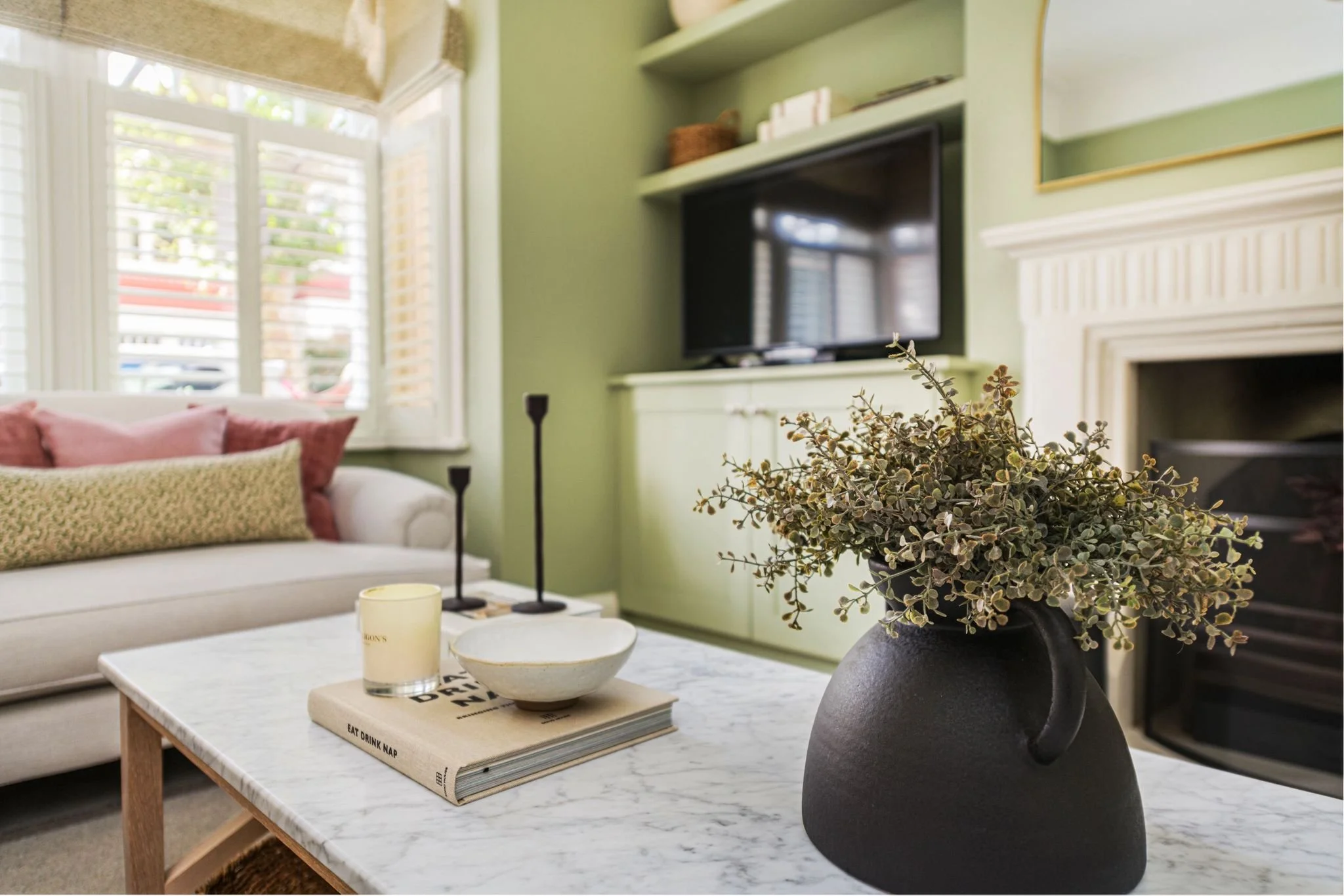 A living room with a white sofa, a marble coffee table with books and a candle, a black vase with green and brown leaves, a wall-mounted TV, and a fireplace, with large windows and green walls.