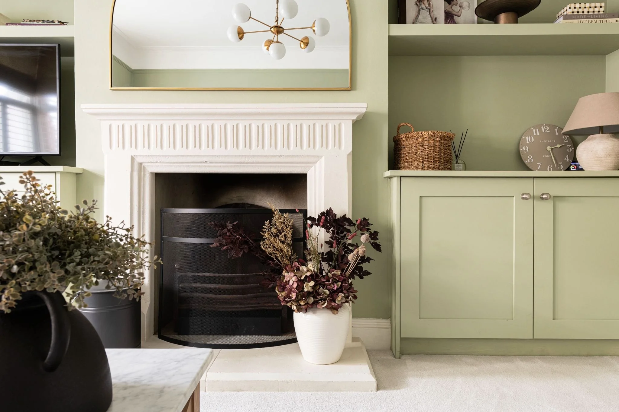 Living room with a fireplace, a green painted wall, a large mirror, a TV, a wicker basket, a clock, and decorative plants and furniture.