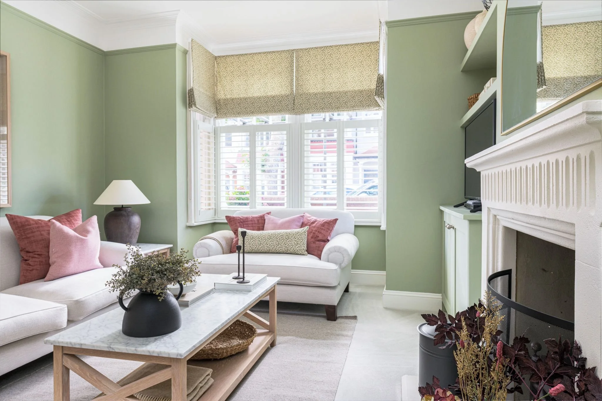 Living room with light green walls, large bay window with white shutters and beige Roman shades, white sofa with pink and beige pillows, black vase with greenery on marble coffee table, white fireplace with mantel, TV on mantel, decorative items abov