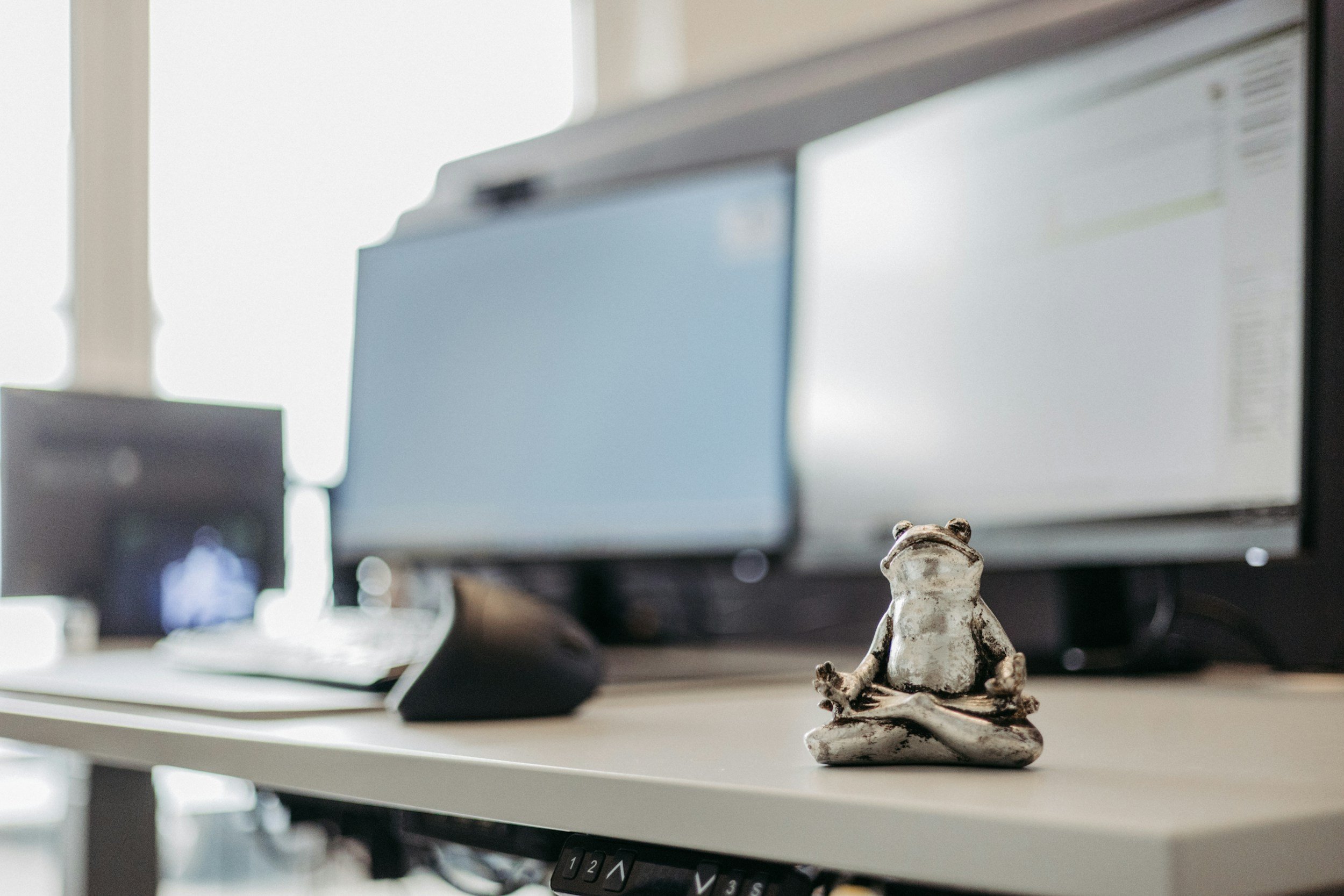 A small ceramic frog figurine sitting on a white desk in front of computers and monitors in an office setting.