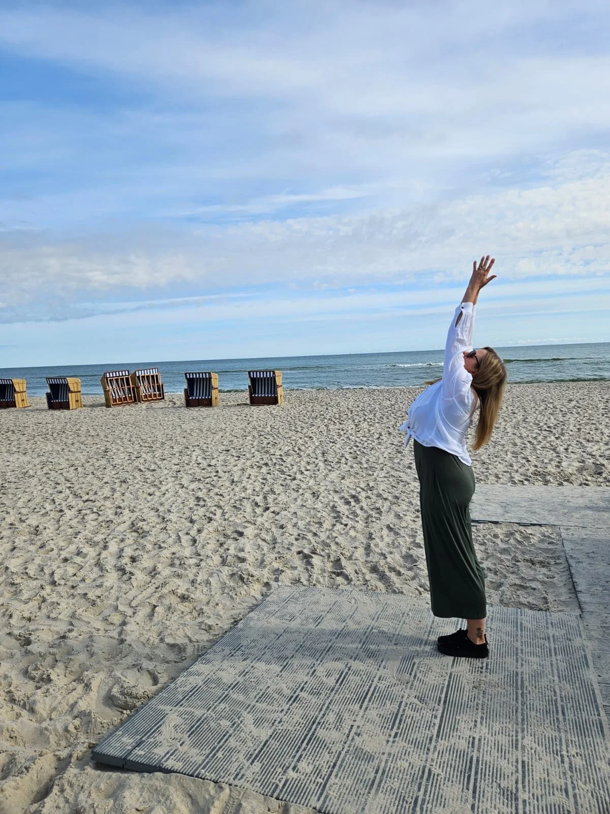 Woman practicing yoga on a mat at the beach with sand, ocean, and blue sky in the background.