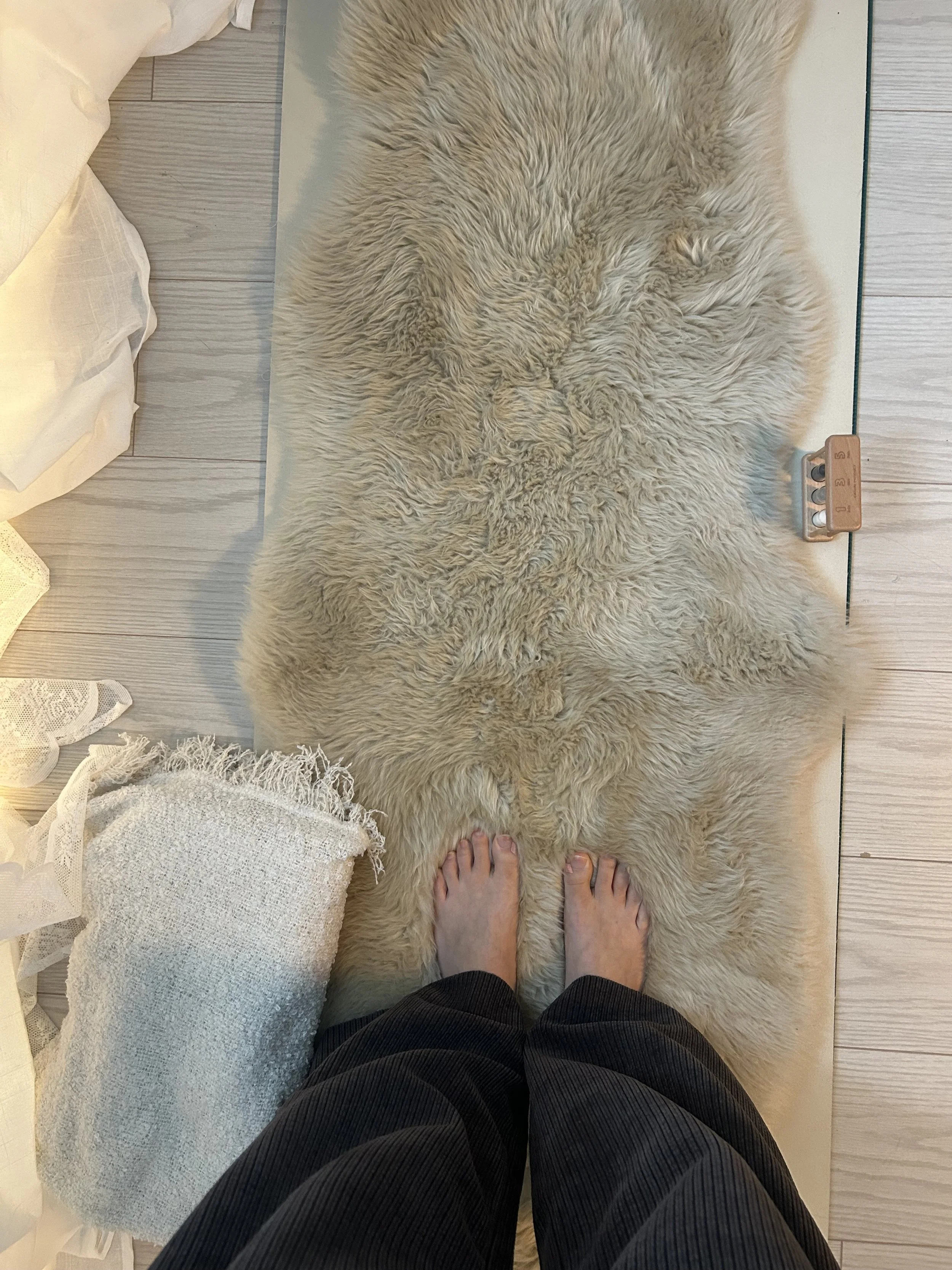 Person standing on a beige shaggy rug on a light wood floor, with bare feet and black pants, with a pillow with a white textured cover and lace trim nearby.