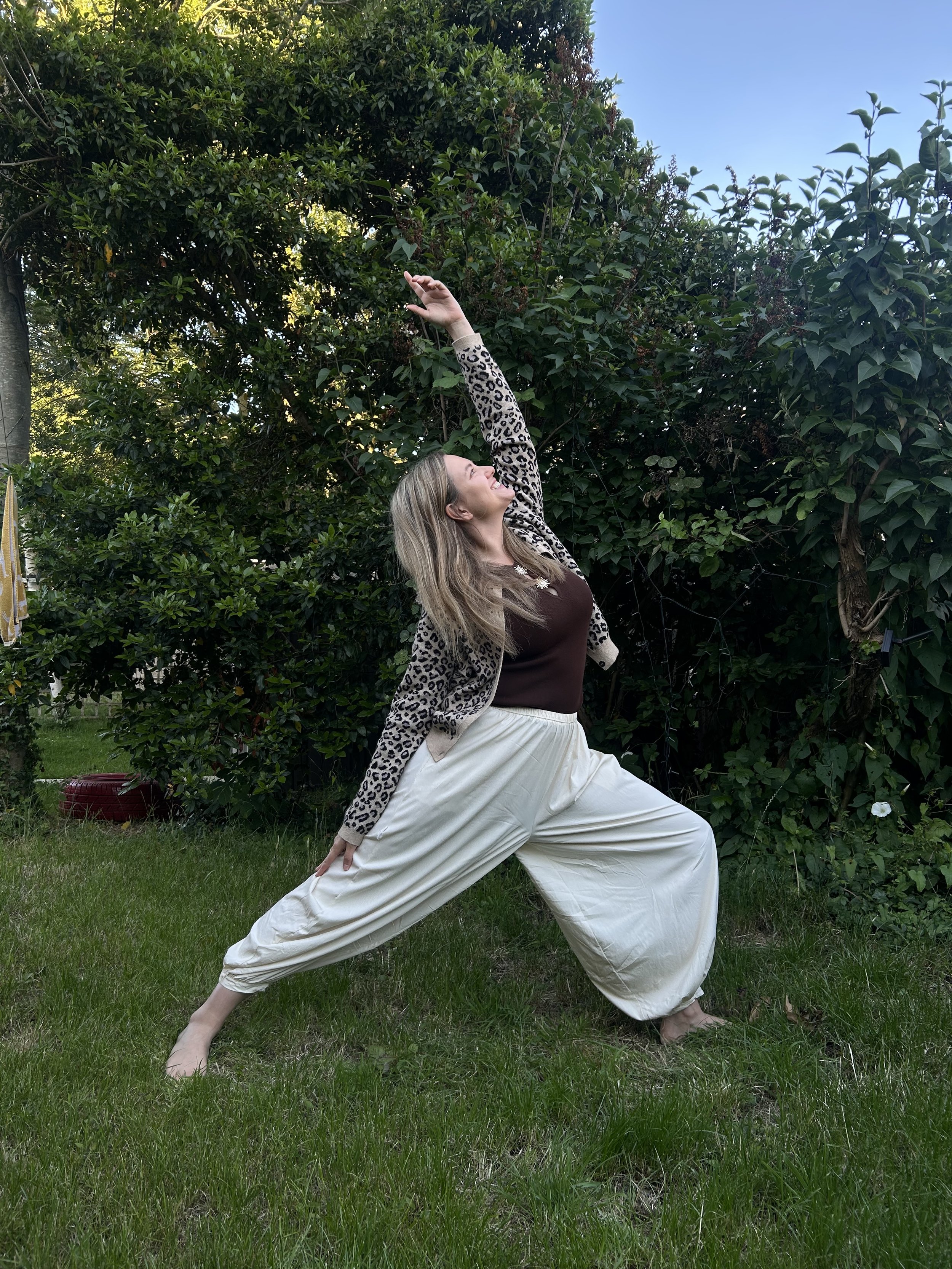 A woman in a yoga pose outdoors on a grassy lawn, wearing a leopard print cardigan, brown top, and white wide-leg pants, with a background of lush green trees and blue sky.