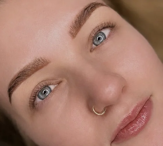 Close-up van een vrouw met blauwe ogen, getinte wenkbrauwen, een neusring, en lichte make-up.