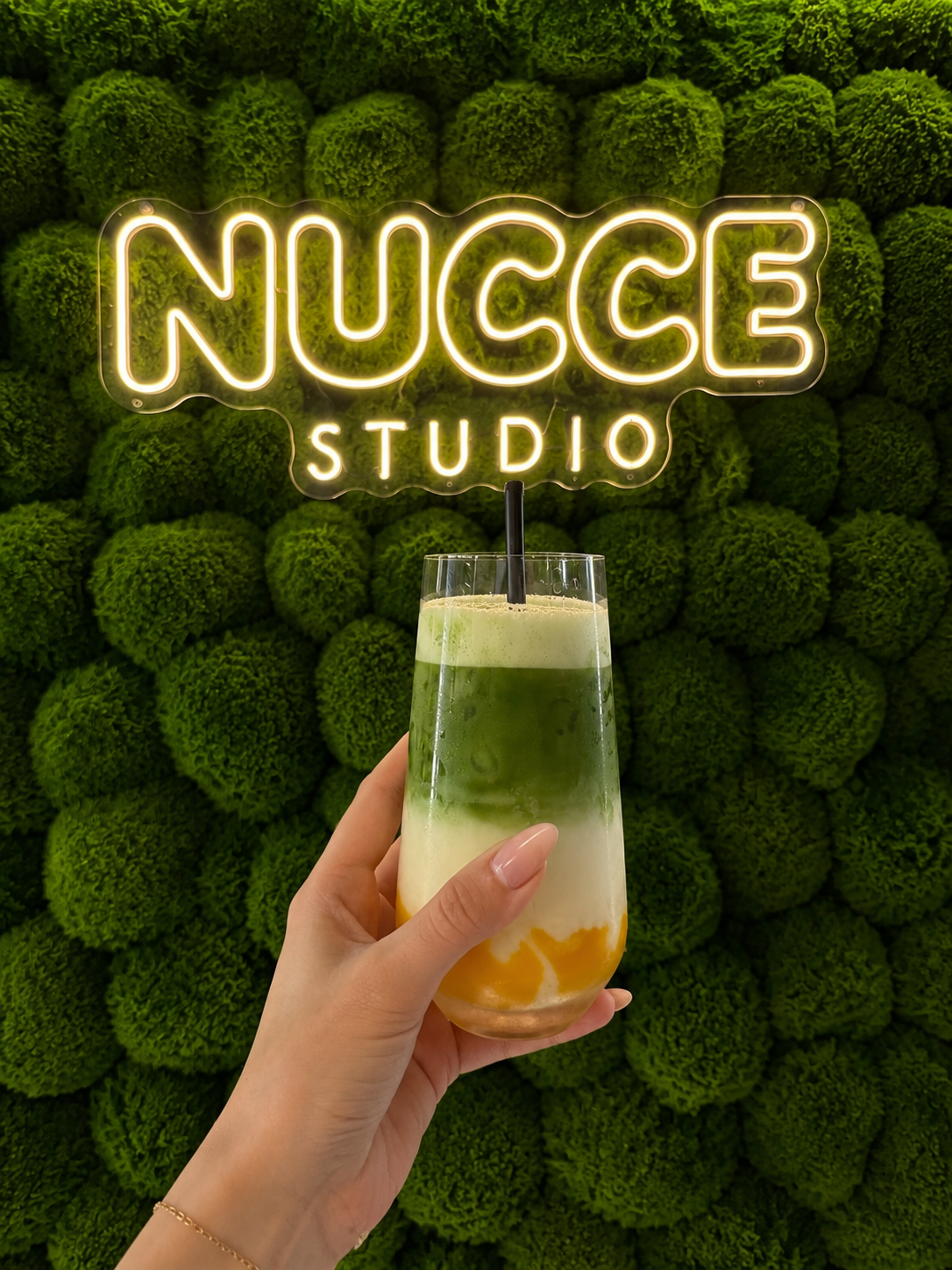 Een hand die een glas met een groen-wit-oranje drankje vasthoudt, op de achtergrond een groengroen muur met een neonbord dat zegt 'NUCCE STUDIO'.