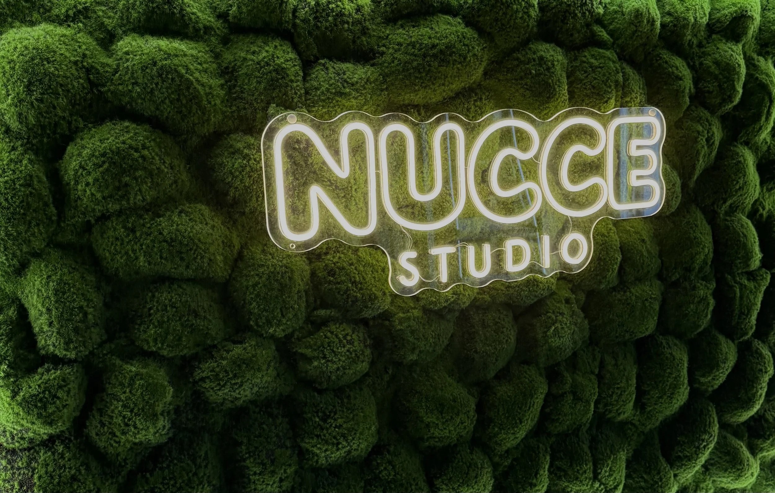 Neonlichtbord met de tekst 'NUAGE STUDIO' op een muur bedekt met groene mosballen.