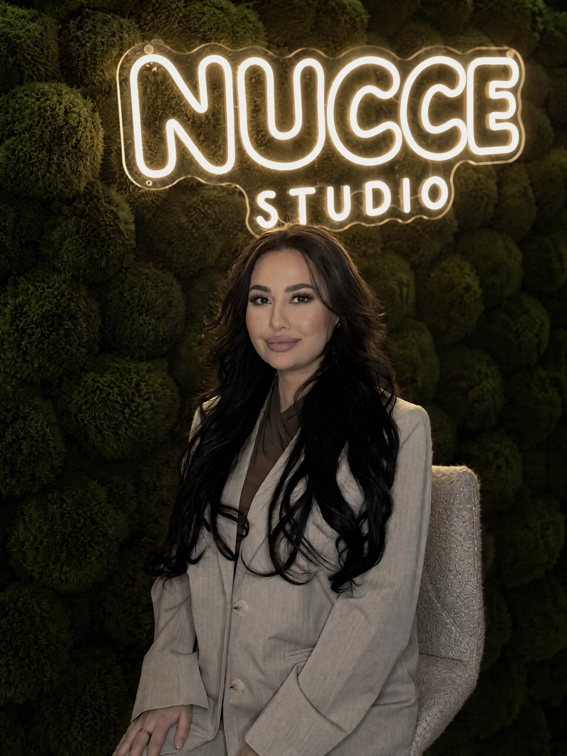 Portret van een vrouw met lang zwart haar, gekleed in een beige broekpak, zittend op een stoel, met een groene, met mos bedekte achtergrond en een neonlichtbord dat zegt 'NUCCE STUDIO'.