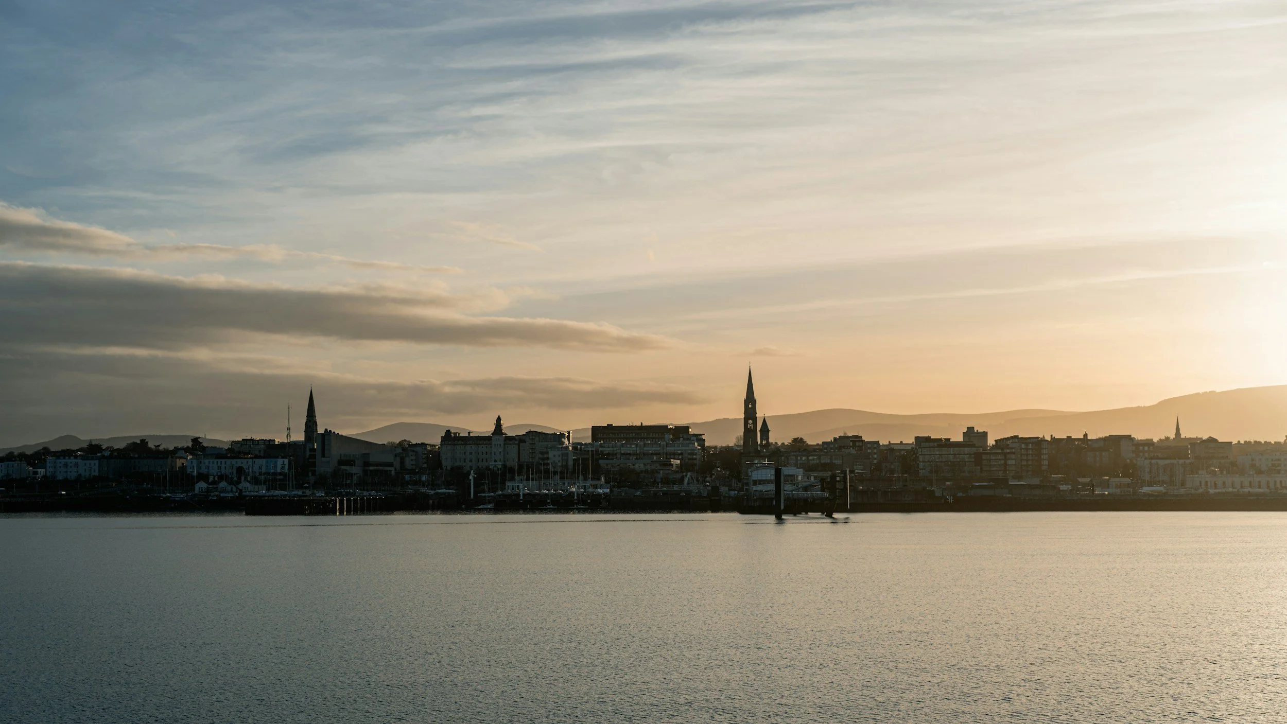 A city skyline viewed from across a body of water during sunset, with several church steeples and buildings, and mountains in the background.