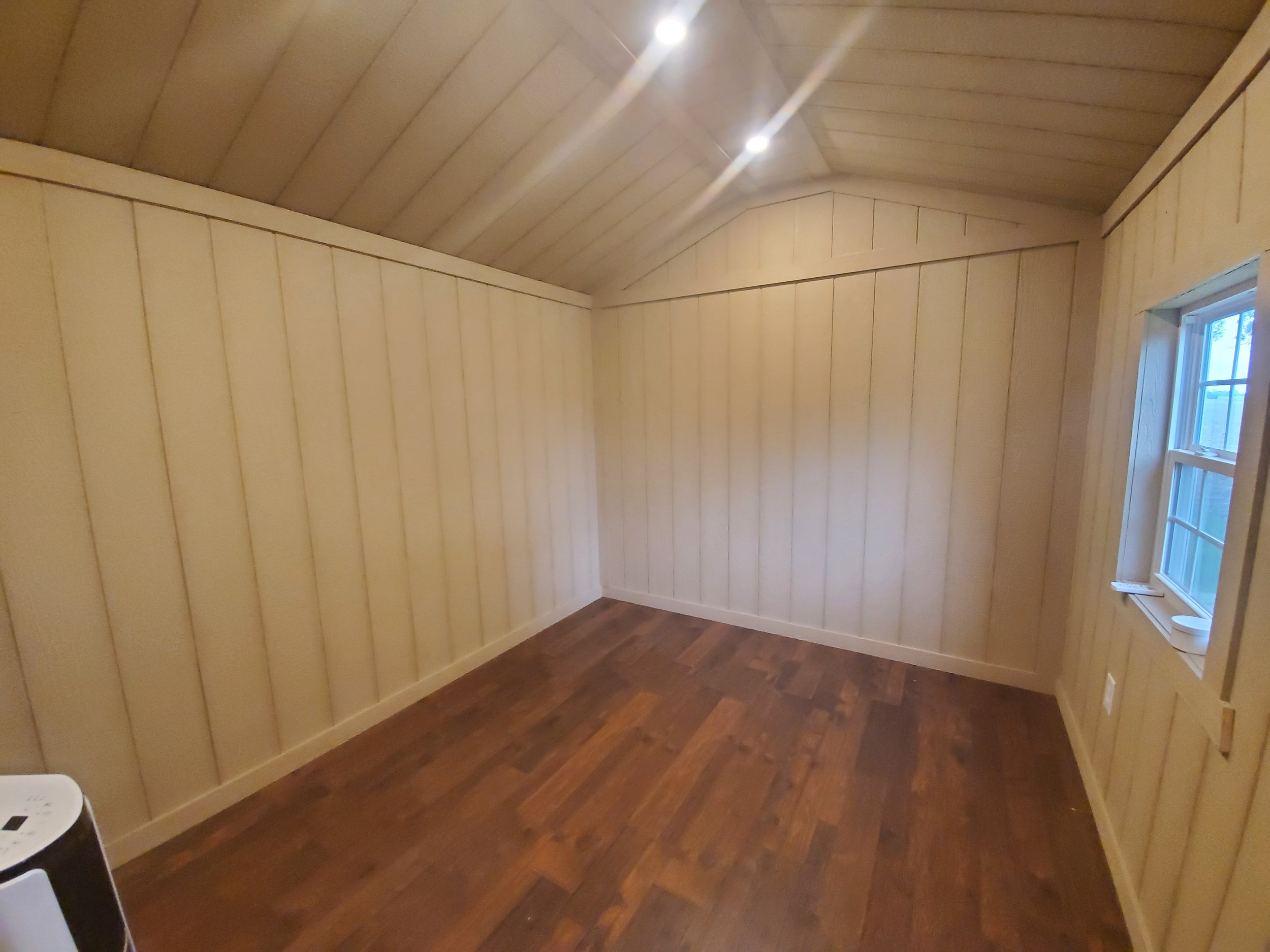 Empty room with light wood panel walls and dark hardwood flooring, small window on the right wall, ceiling lights turned on.