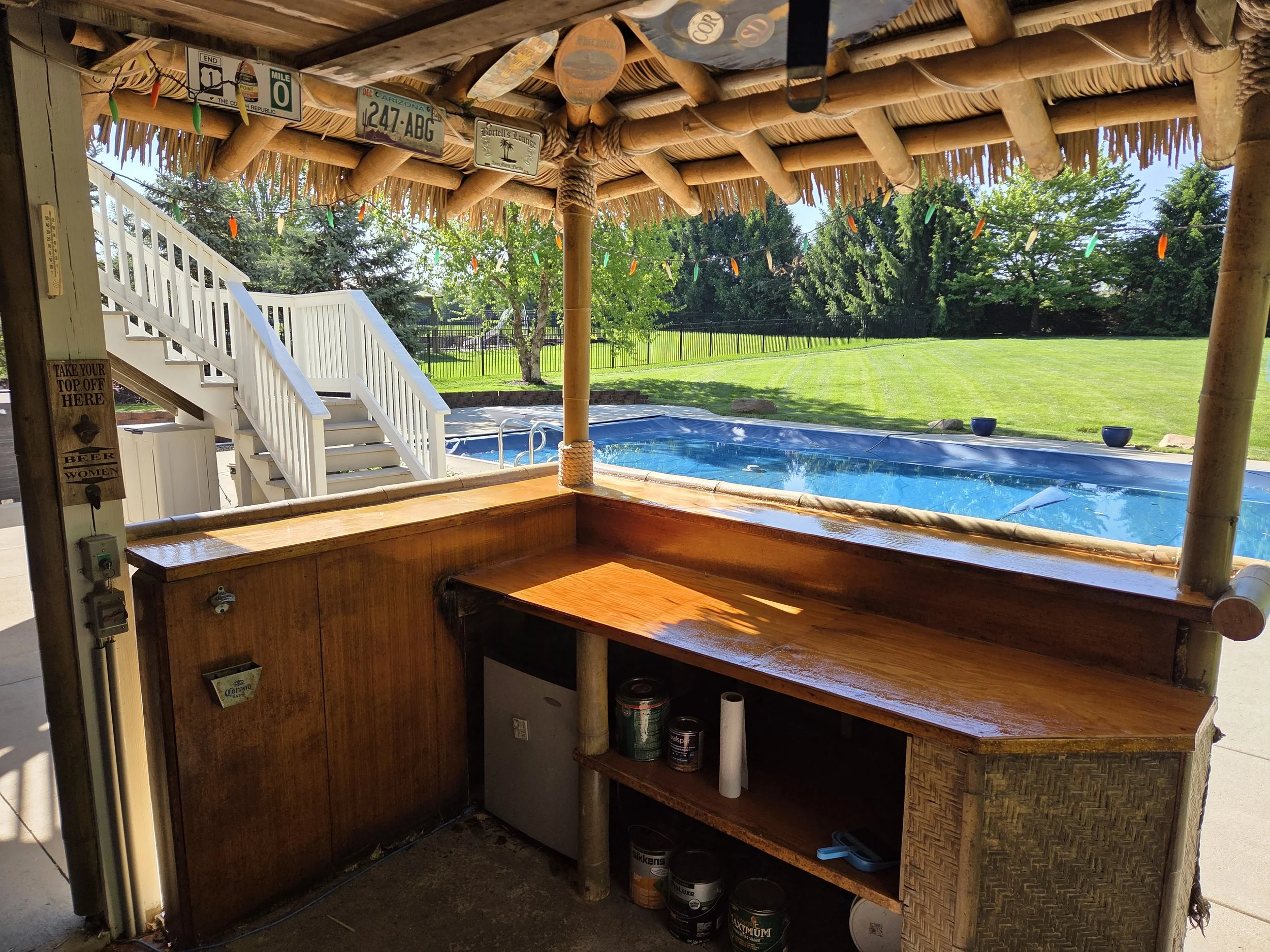 View from inside a tiki hut overlooking a backyard with green grass, trees, and a black fence. The hut has a thatched roof decorated with colorful string lights, and a small pool below.