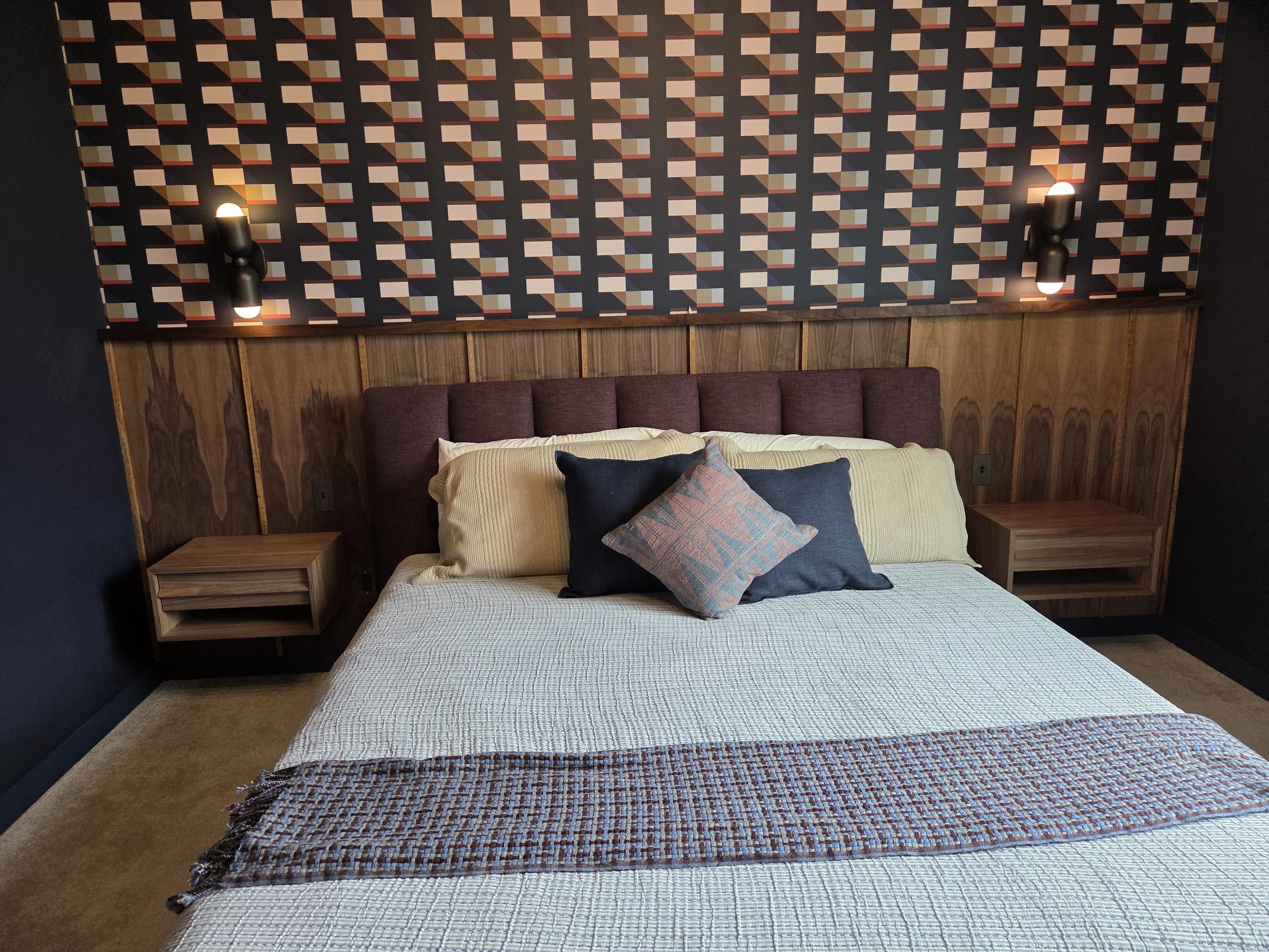 A bedroom featuring a bed with a beige quilt, dark throw pillows, and a colorful woven blanket at the foot. Behind the bed is a wooden headboard with a geometric patterned wallpaper in shades of beige, pink, green, and black. Two black wall-mounted l