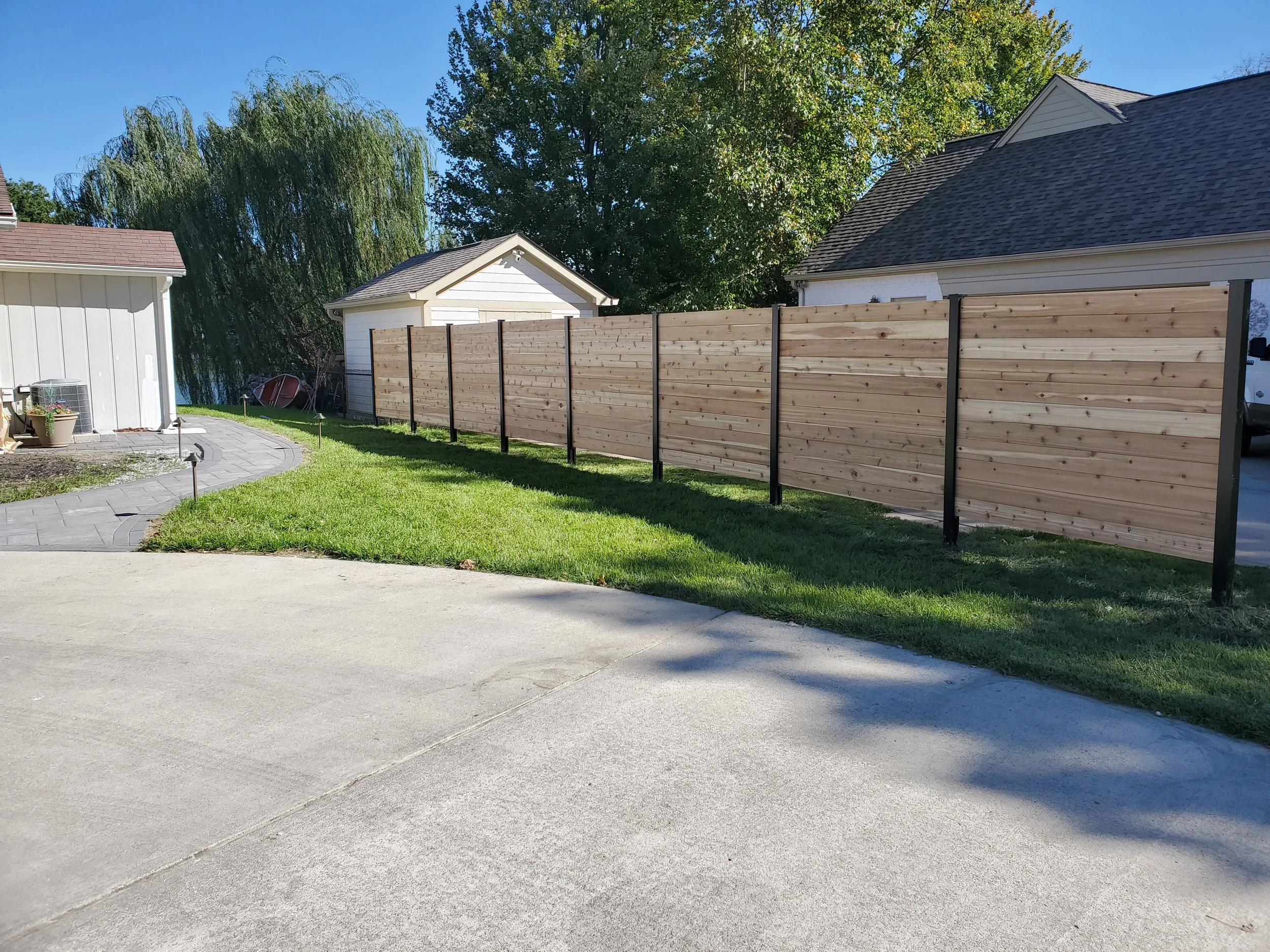 A backyard with a wooden privacy fence, small accessory structures, a paved walkway, and a well-maintained grassy area. Large trees and a house with a shingled roof are visible in the background.