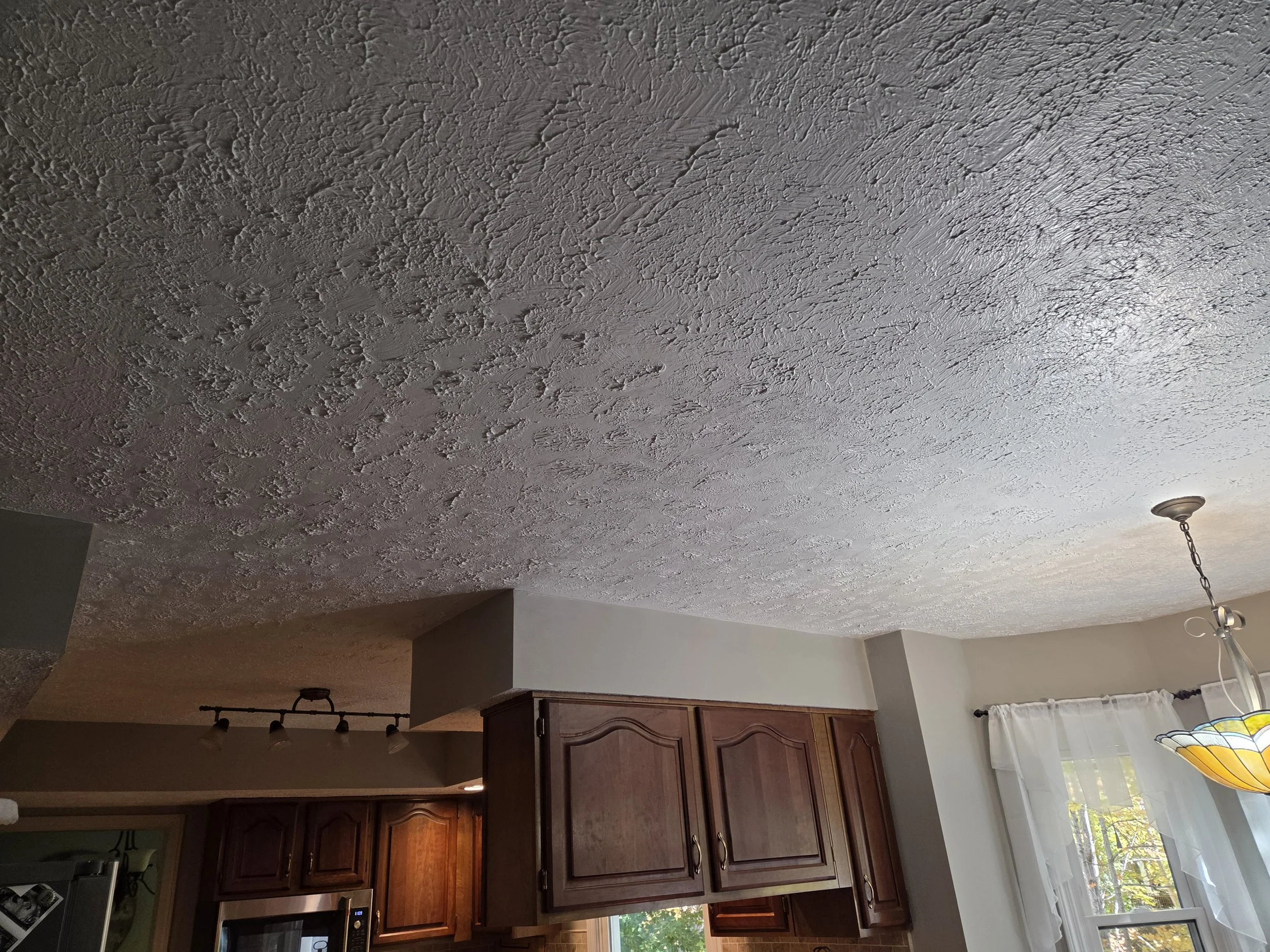 Ceiling with textured, bumpy finish in a living area, kitchen cabinets in the background, window with white curtains, and a pendant light hanging from the ceiling.
