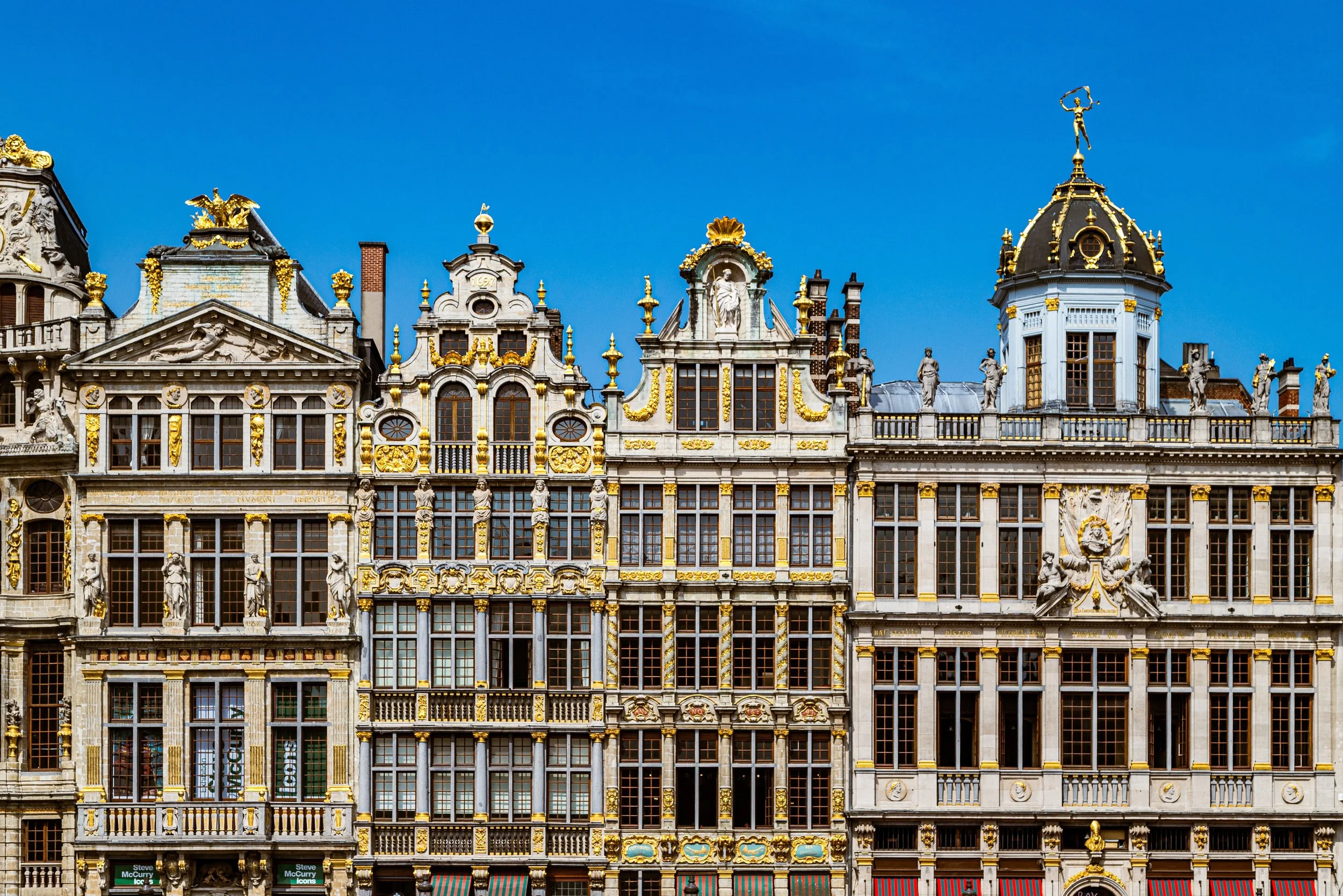 Facade of ornate historic buildings with intricate architectural details, sculptures, and gold accents under a clear blue sky.