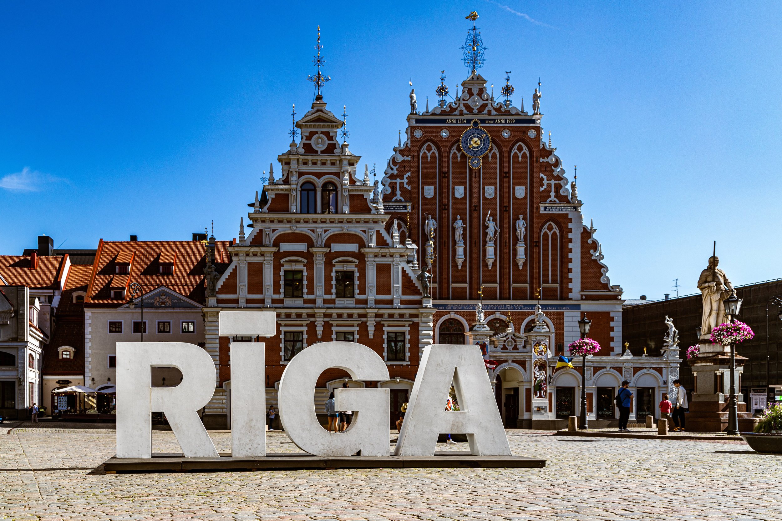 The image shows the historic Town Hall Square with the Riga Town Hall and the House of the Blackheads in Riga, Latvia. There is a large white sign spelling "RIGA" in front of the buildings, and a few people are walking around. The sky is clear and blue.