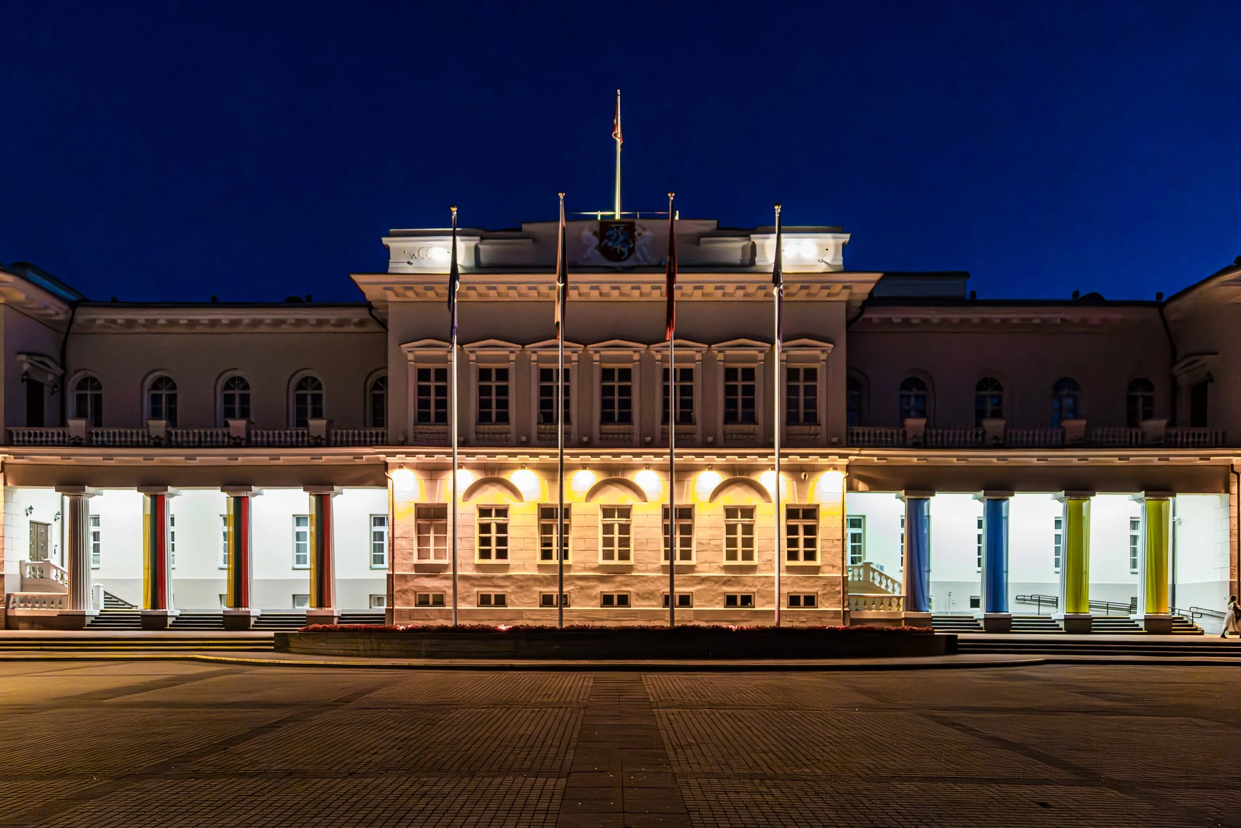 Night view of a grand, illuminated historic building with flags in front and steps leading up to the entrance.