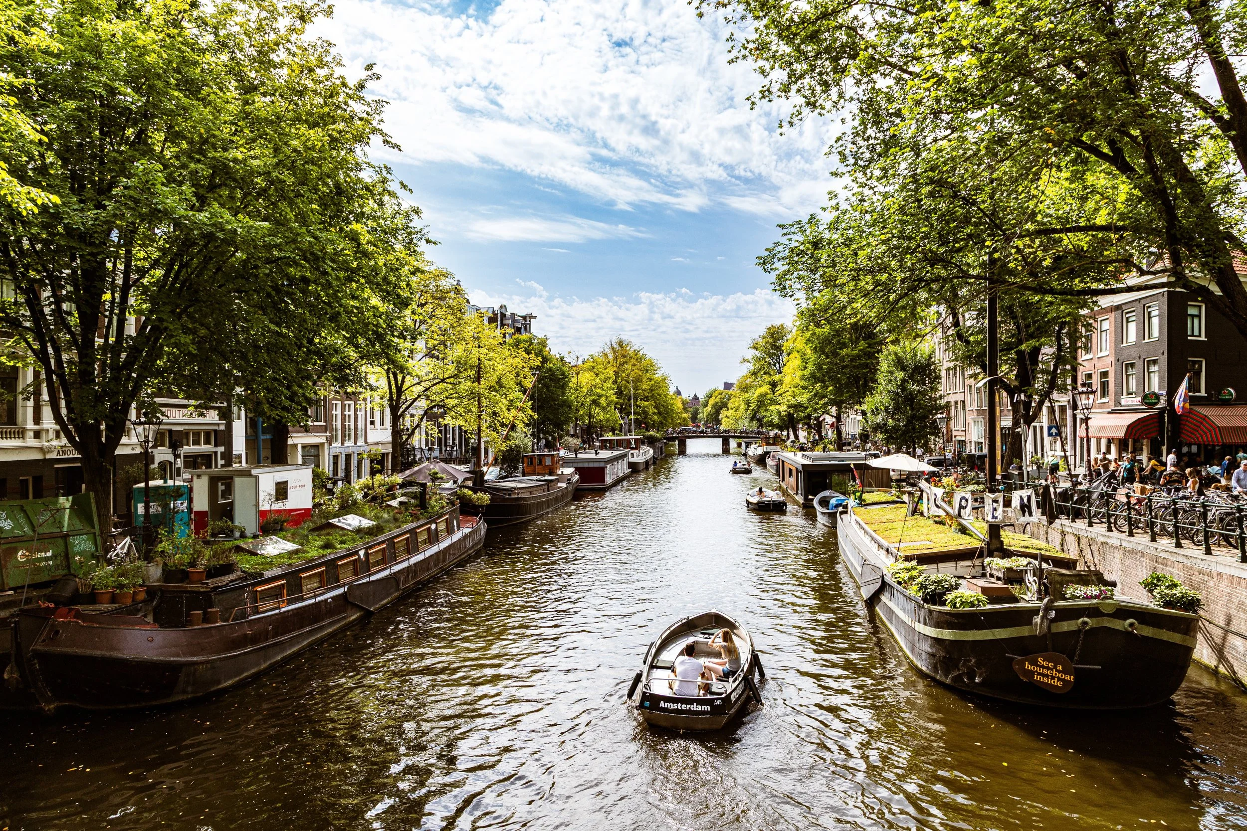 A canal scene in Amsterdam with boats on the water, houseboats along the sides, and trees lining the canal. There are people on a boat and on the sidewalk, with a bridge in the distance and a partly cloudy sky overhead.