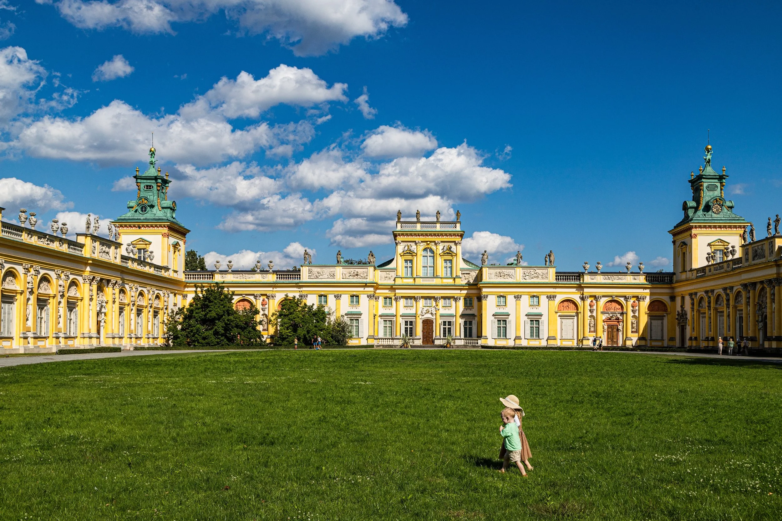 A large yellow and white baroque-style palace with green roofs and towers, set against a blue sky with scattered clouds. In front, there is a grassy lawn with two young children, one wearing a sunhat, playing near each other.