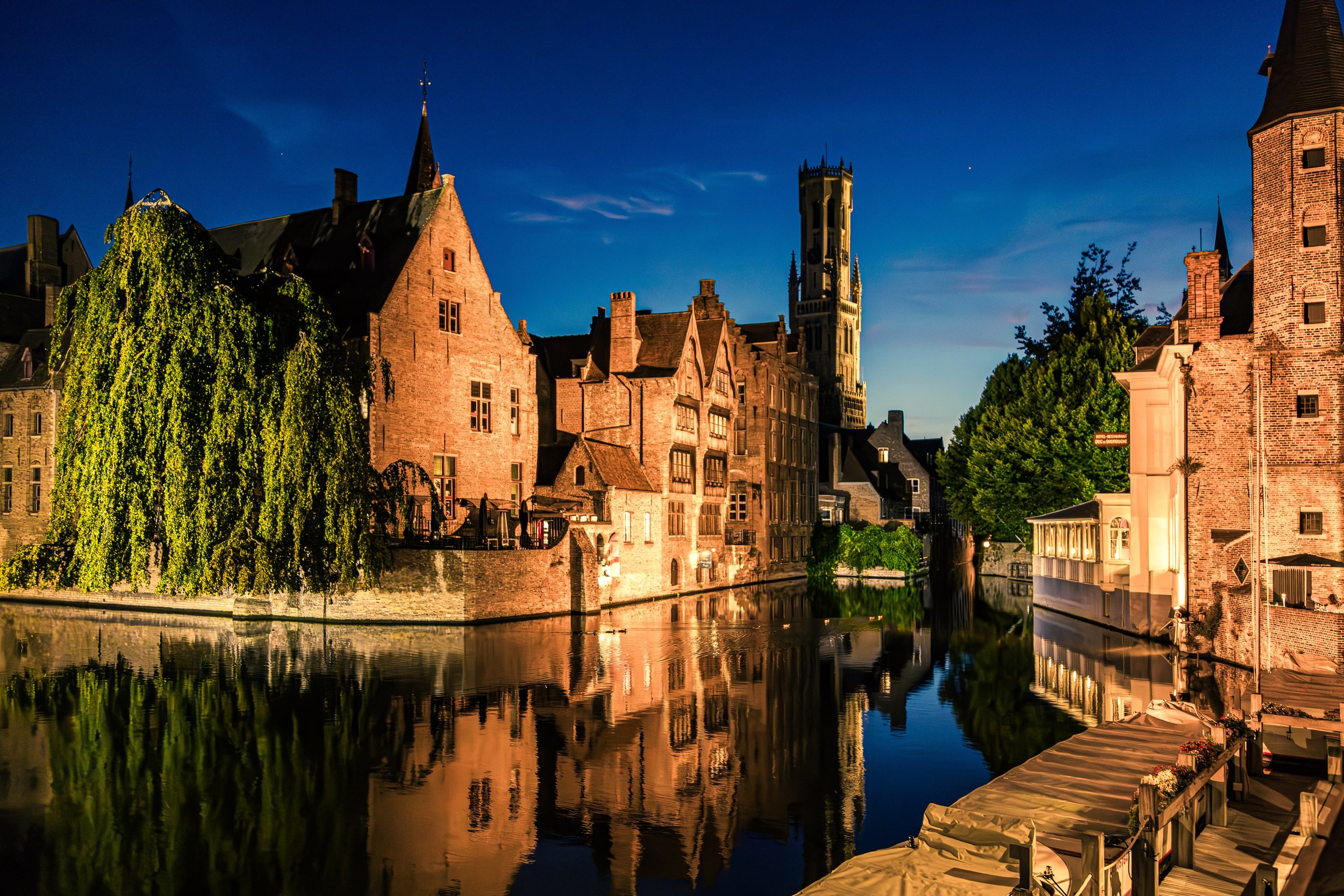 Nighttime view of historic European buildings with a canal, reflecting the illuminated structures and a blue sky.