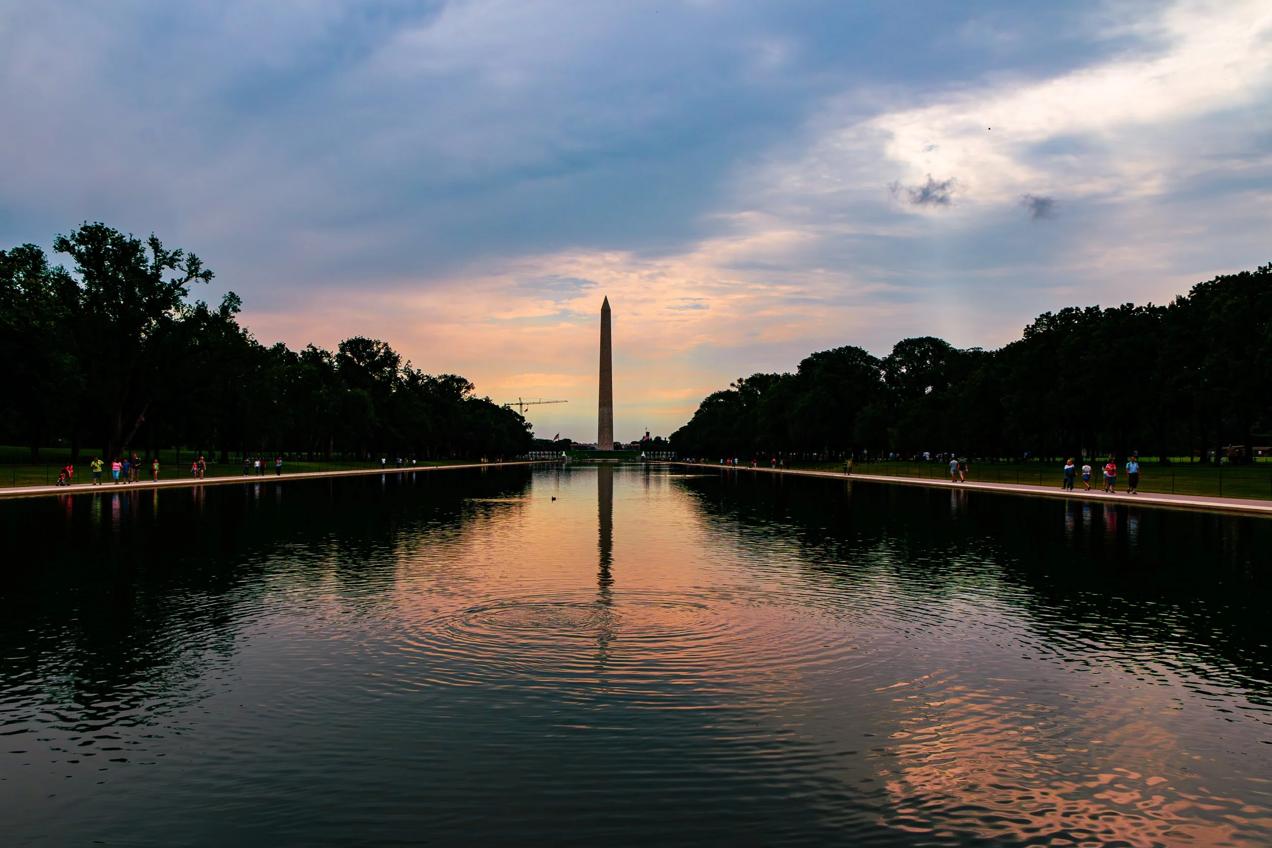 View of the Washington Monument reflected in the Reflecting Pool at sunset with a sky filled with clouds and people walking along the pool on both sides.
