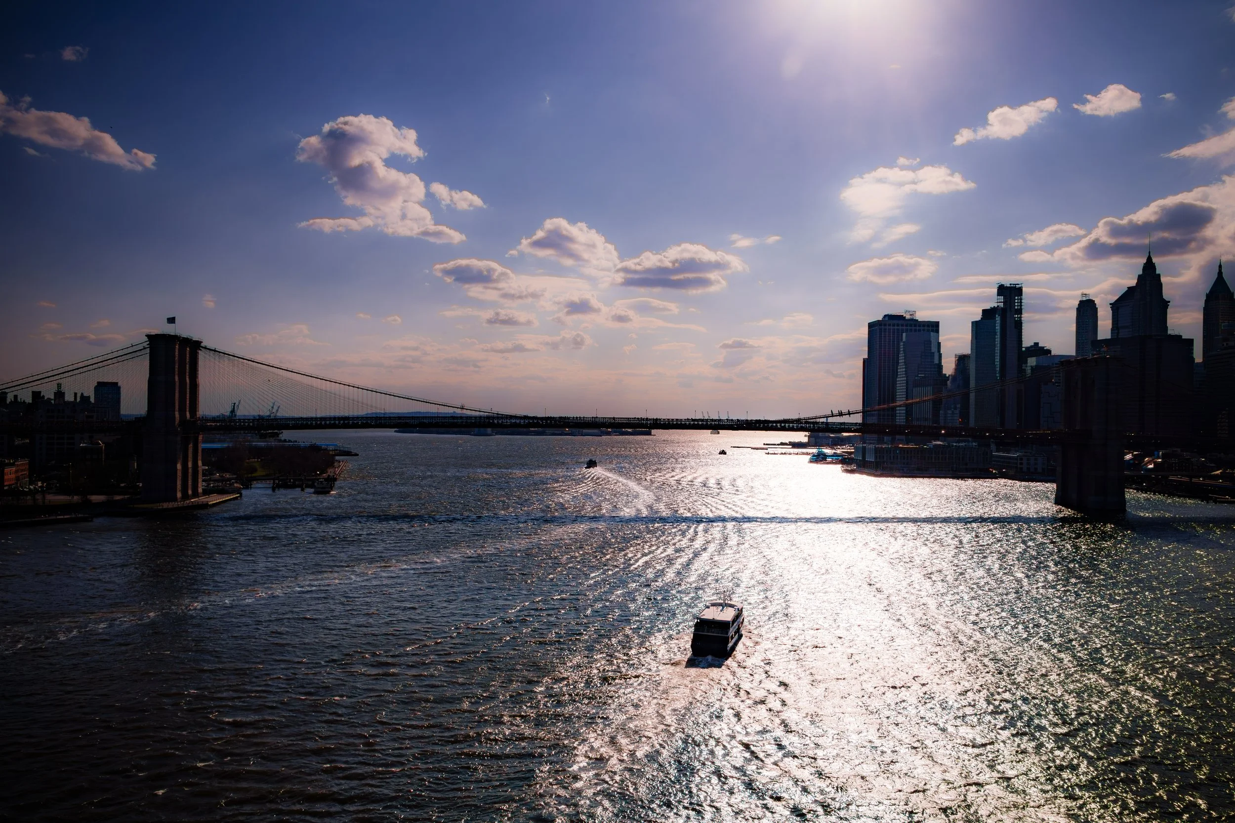 View of the Manhattan Bridge and skyline in New York City with a boat on the water at sunset.