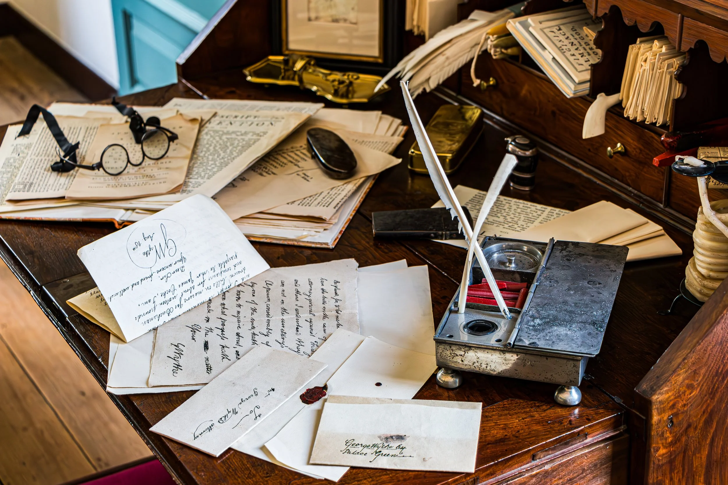 A vintage wooden desk cluttered with papers, handwritten letters, a pair of round glasses, a black seal, and an antique ink stamp with a feather quill. There is also an open file drawer with more papers and a prince of a paper that reads 'Mr. G.'