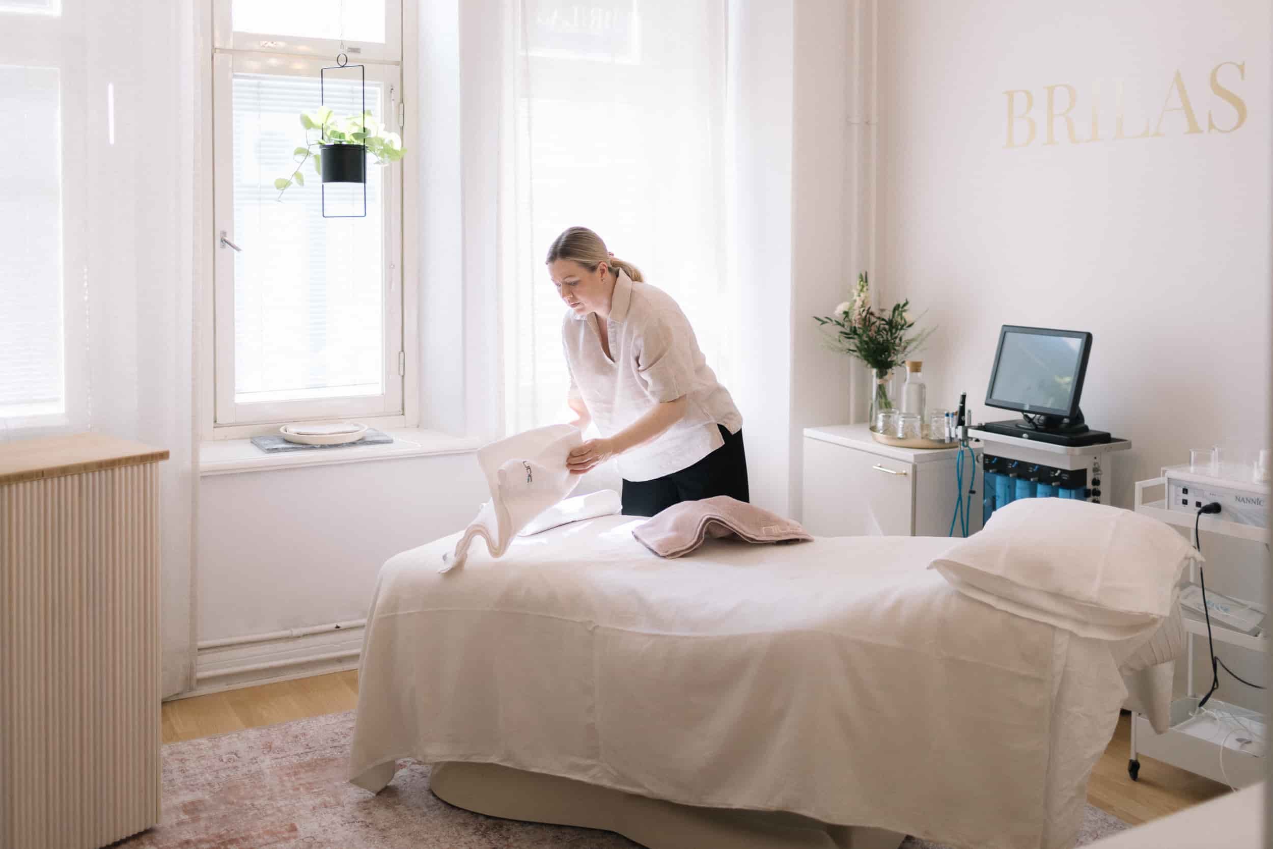 Kosmetologi petaa vuoteen pedin kauneushoitolassa, jossa on suuri ikkuna ja Hydrafacial-laite taustalla.