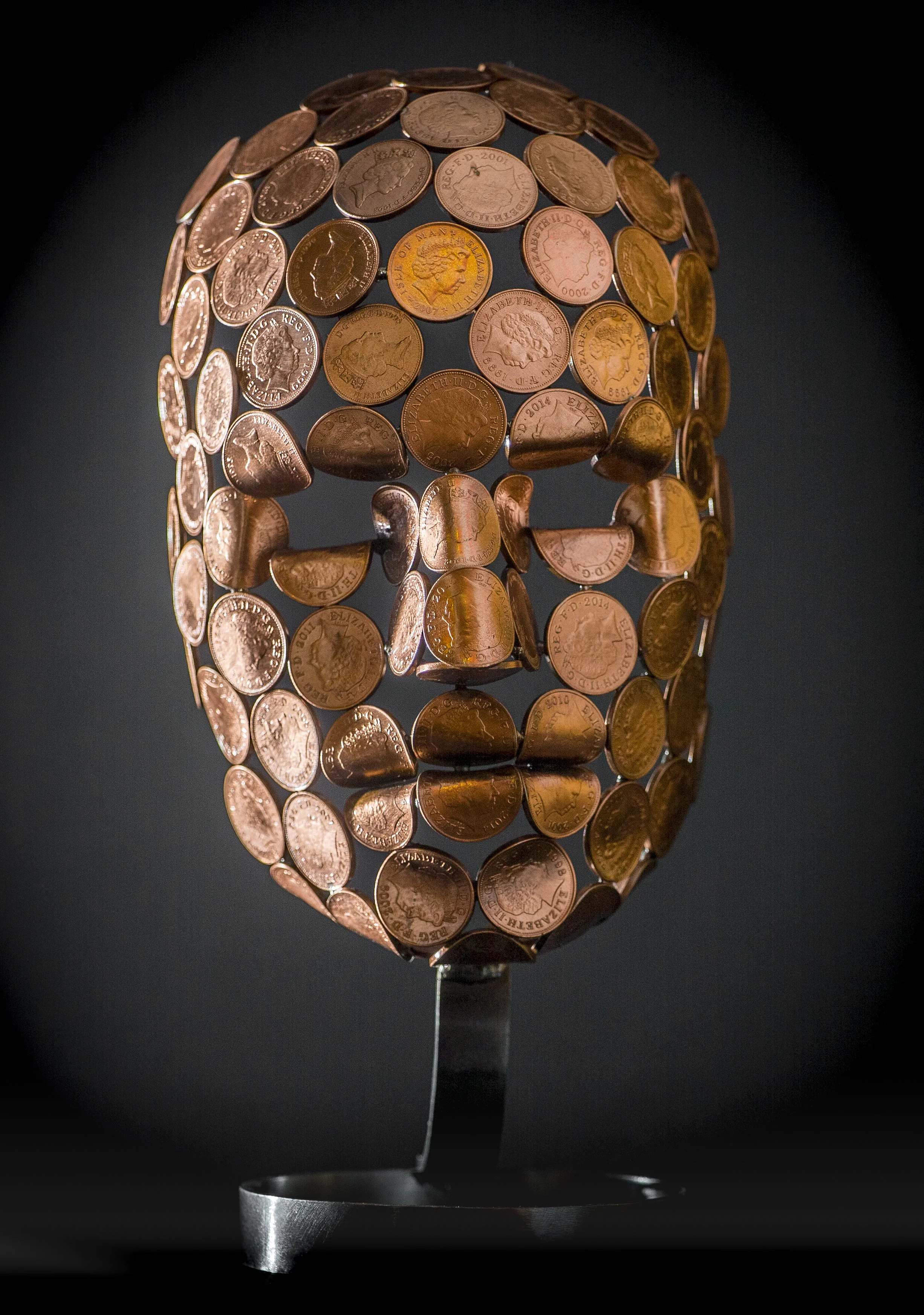 A small face sculpture by Shaun Gagg, made by welding 2p or 1p coins, with some coins folded for greater definition. The sculpture stands on a stylish stainless steel base.