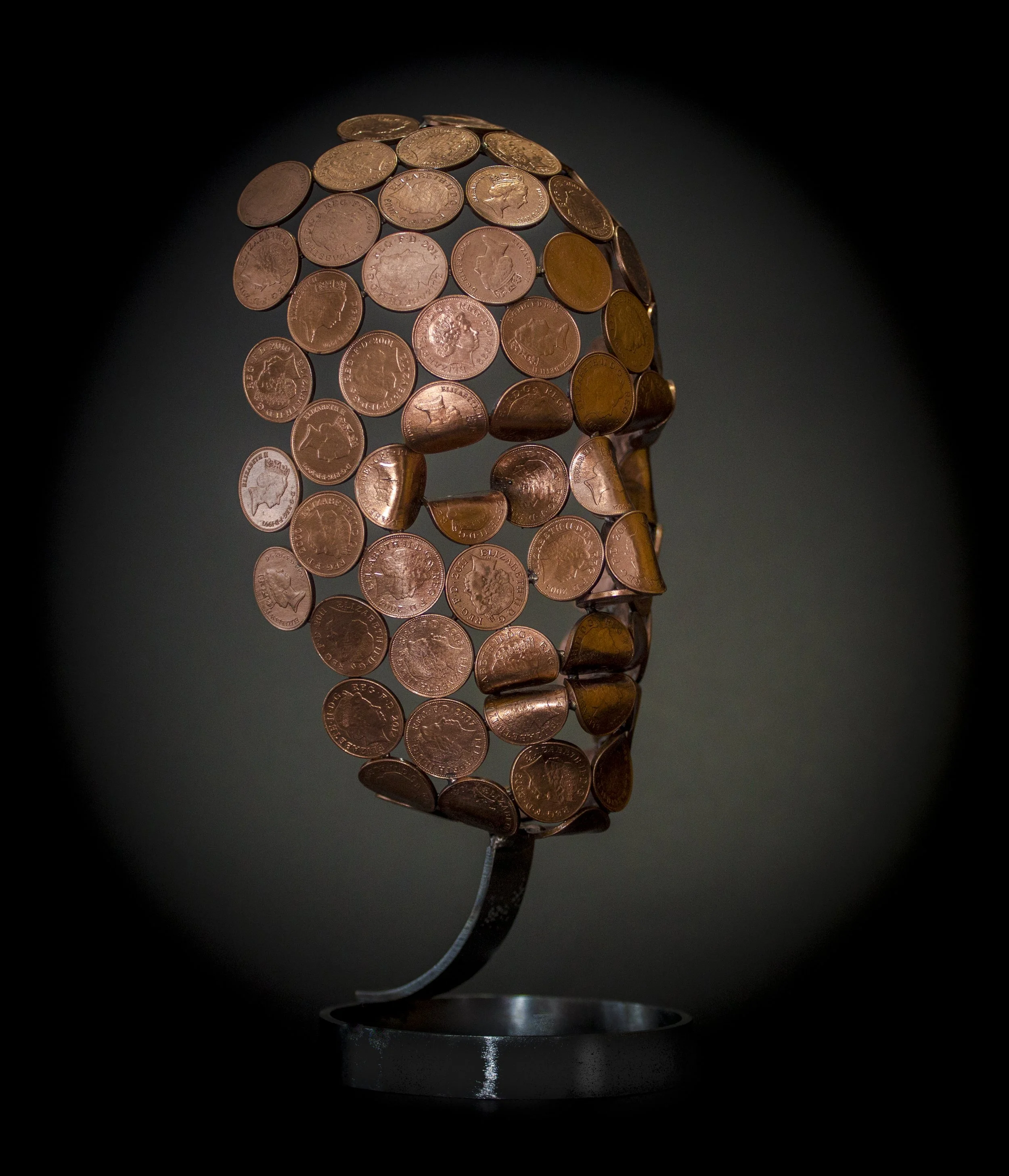 A small face sculpture by Shaun Gagg, made by welding 2p or 1p coins, with some coins folded for greater definition. The sculpture stands on a stylish stainless steel base.