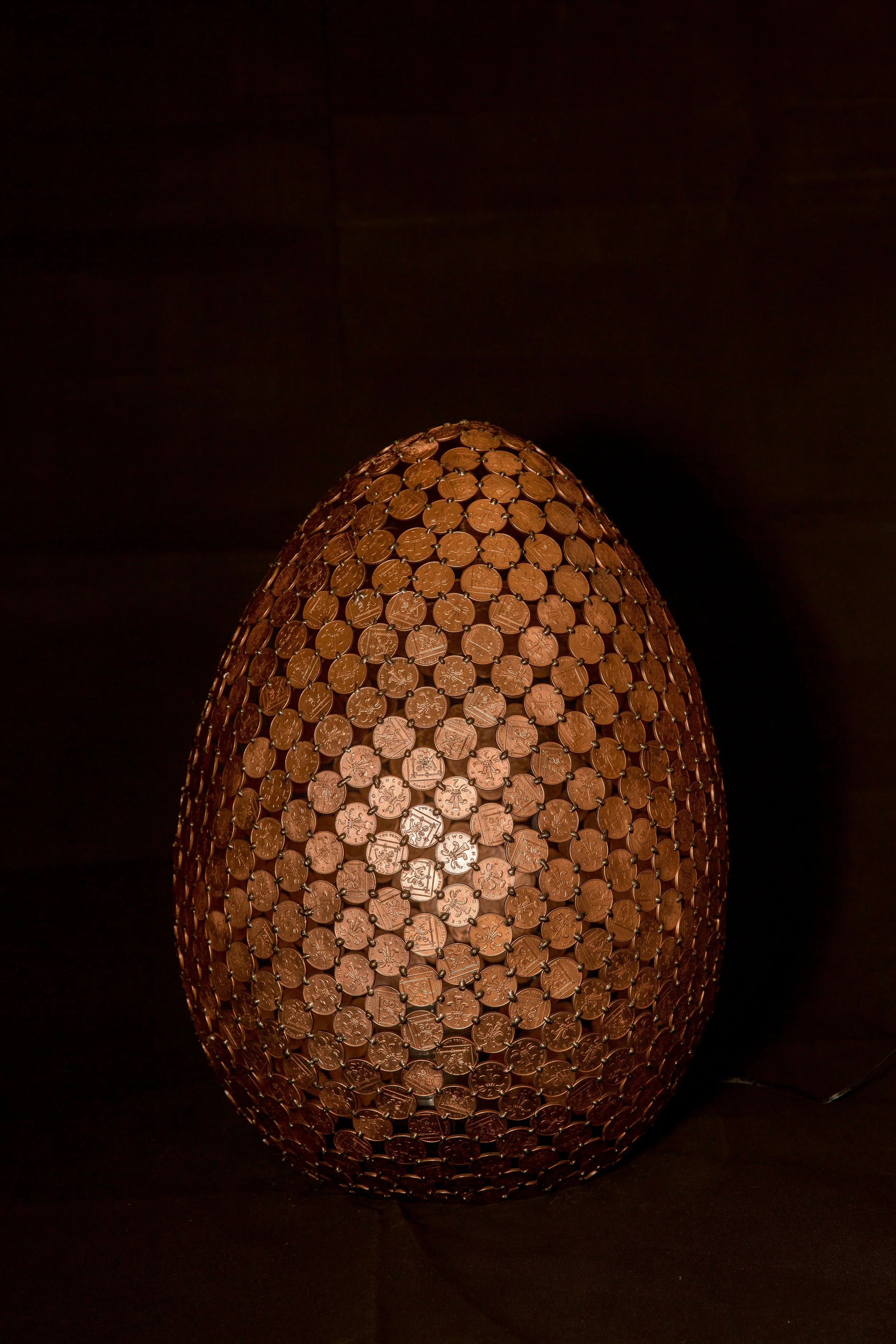 Egg Lamps are large egg-shaped sculptures made from welded 2p coins that emit light with a color-changing controller. Handmade by Shaun Gagg, each piece may vary, and customizations are available.