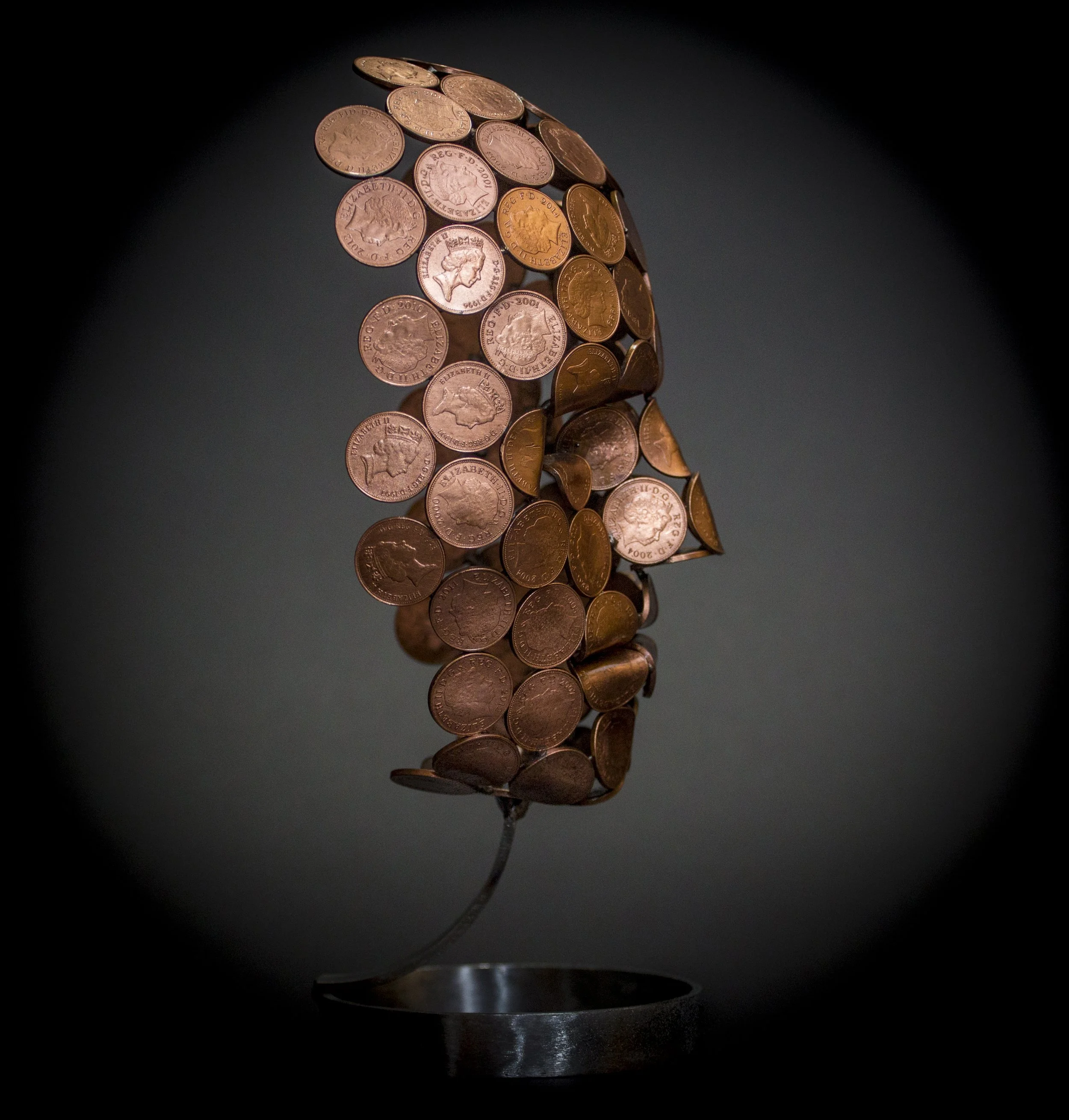 A small face sculpture by Shaun Gagg, made by welding 2p or 1p coins, with some coins folded for greater definition. The sculpture stands on a stylish stainless steel base.
