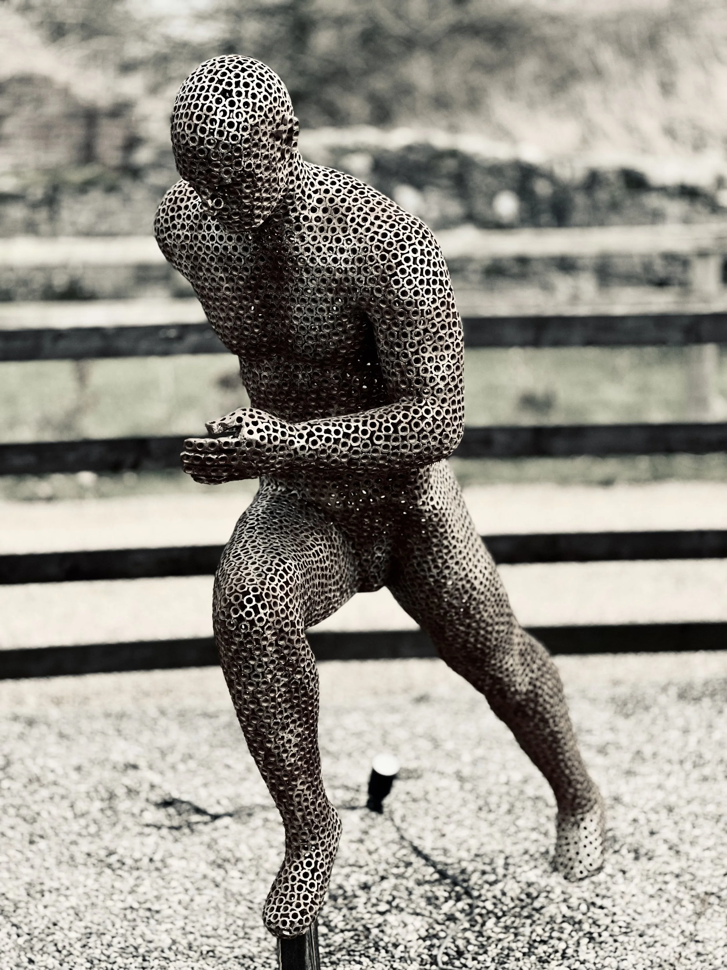 A metal sculpture of a running man, designed by Shaun Gagg in a dynamic pose with one foot lifted off the ground. The figure is made of nuts, creating a semi-transparent, honeycomb-like structure.