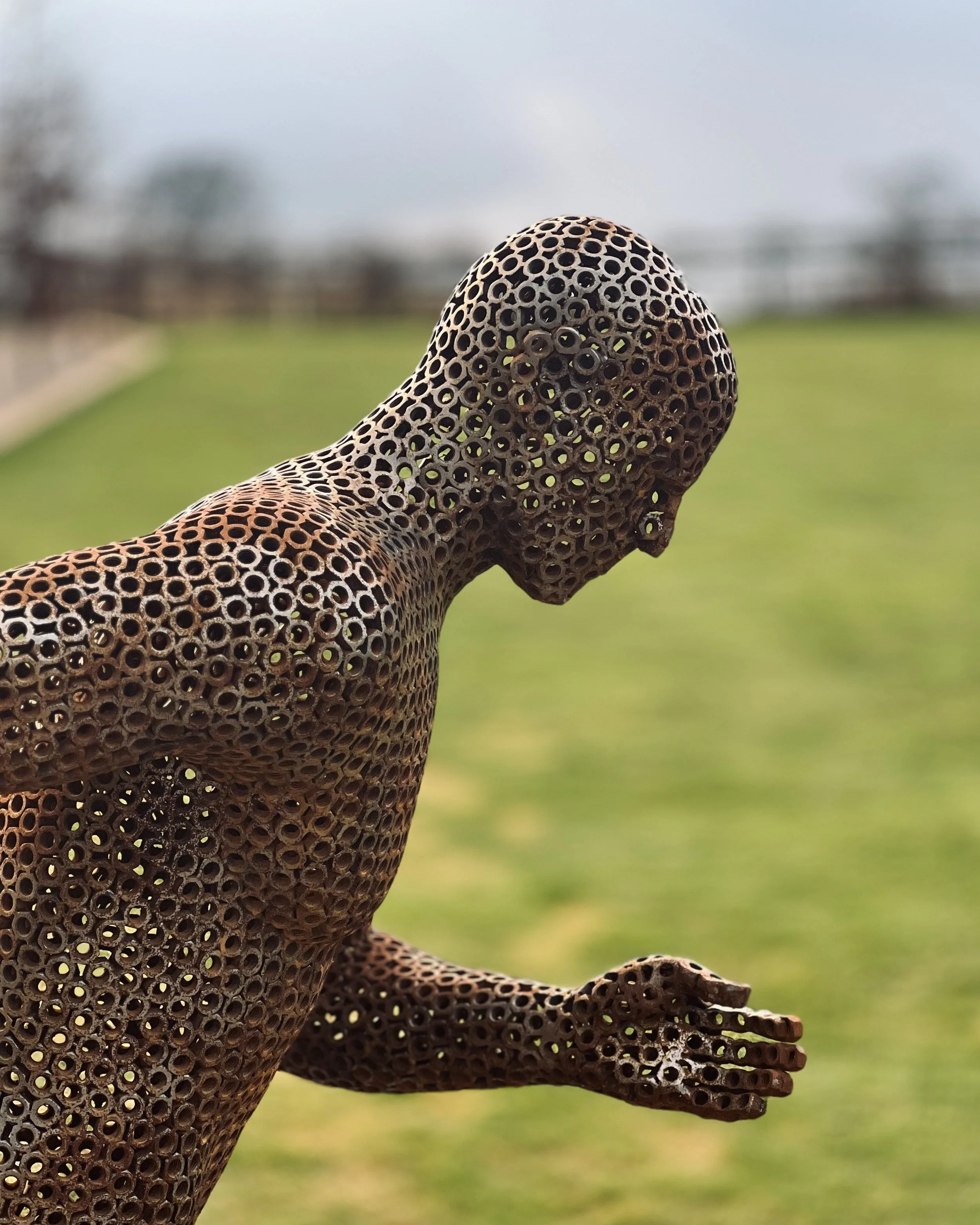 A metal sculpture of a running man, designed by Shaun Gagg in a dynamic pose with one foot lifted off the ground. The figure is made of nuts, creating a semi-transparent, honeycomb-like structure.