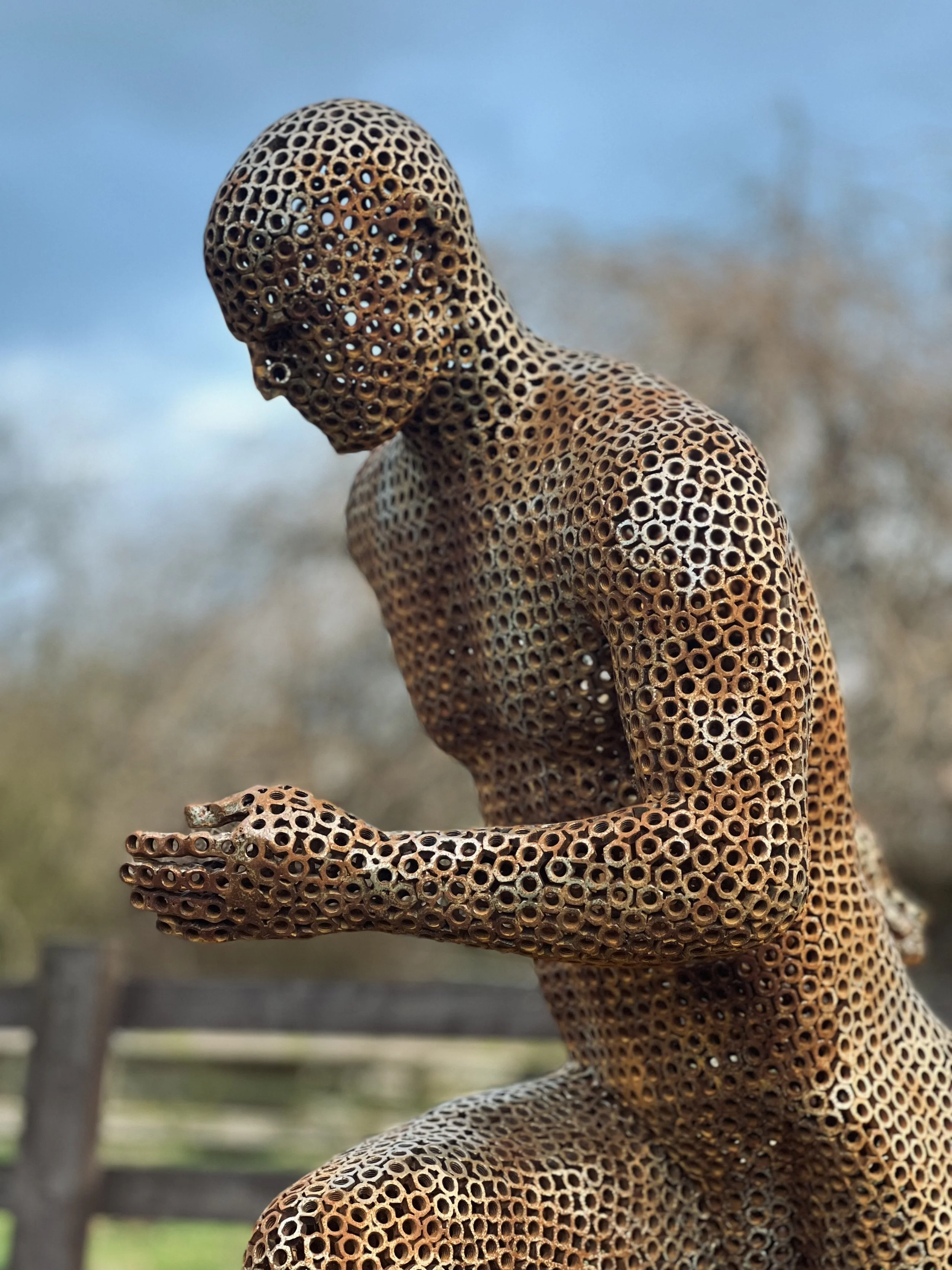 A metal sculpture of a running man, designed by Shaun Gagg in a dynamic pose with one foot lifted off the ground. The figure is made of nuts, creating a semi-transparent, honeycomb-like structure.