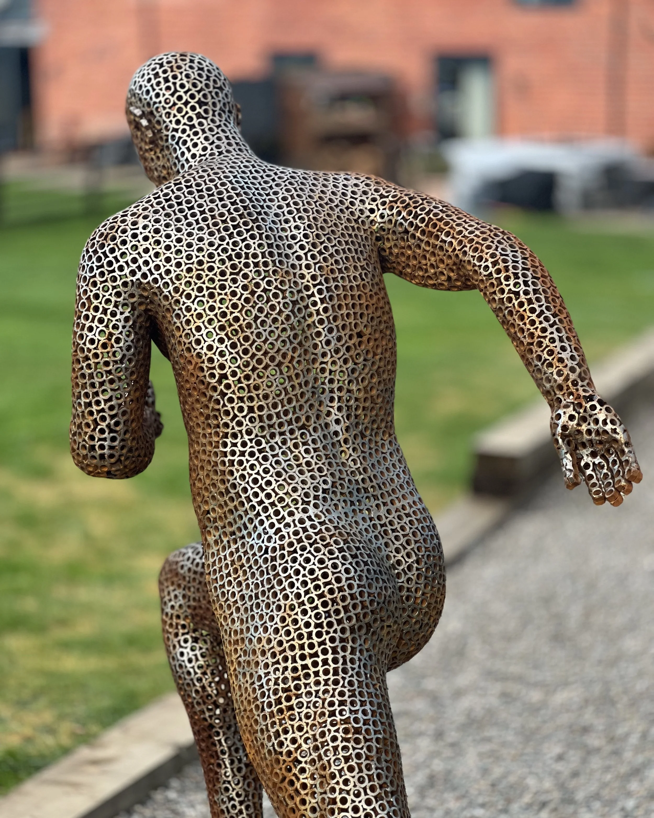 A metal sculpture of a running man, designed by Shaun Gagg in a dynamic pose with one foot lifted off the ground. The figure is made of nuts, creating a semi-transparent, honeycomb-like structure.