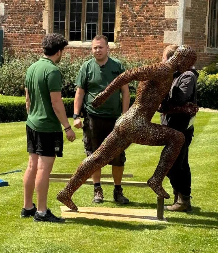 A metal sculpture of a running man, designed by Shaun Gagg in a dynamic pose with one foot lifted off the ground. The figure is made of nuts, creating a semi-transparent, honeycomb-like structure.