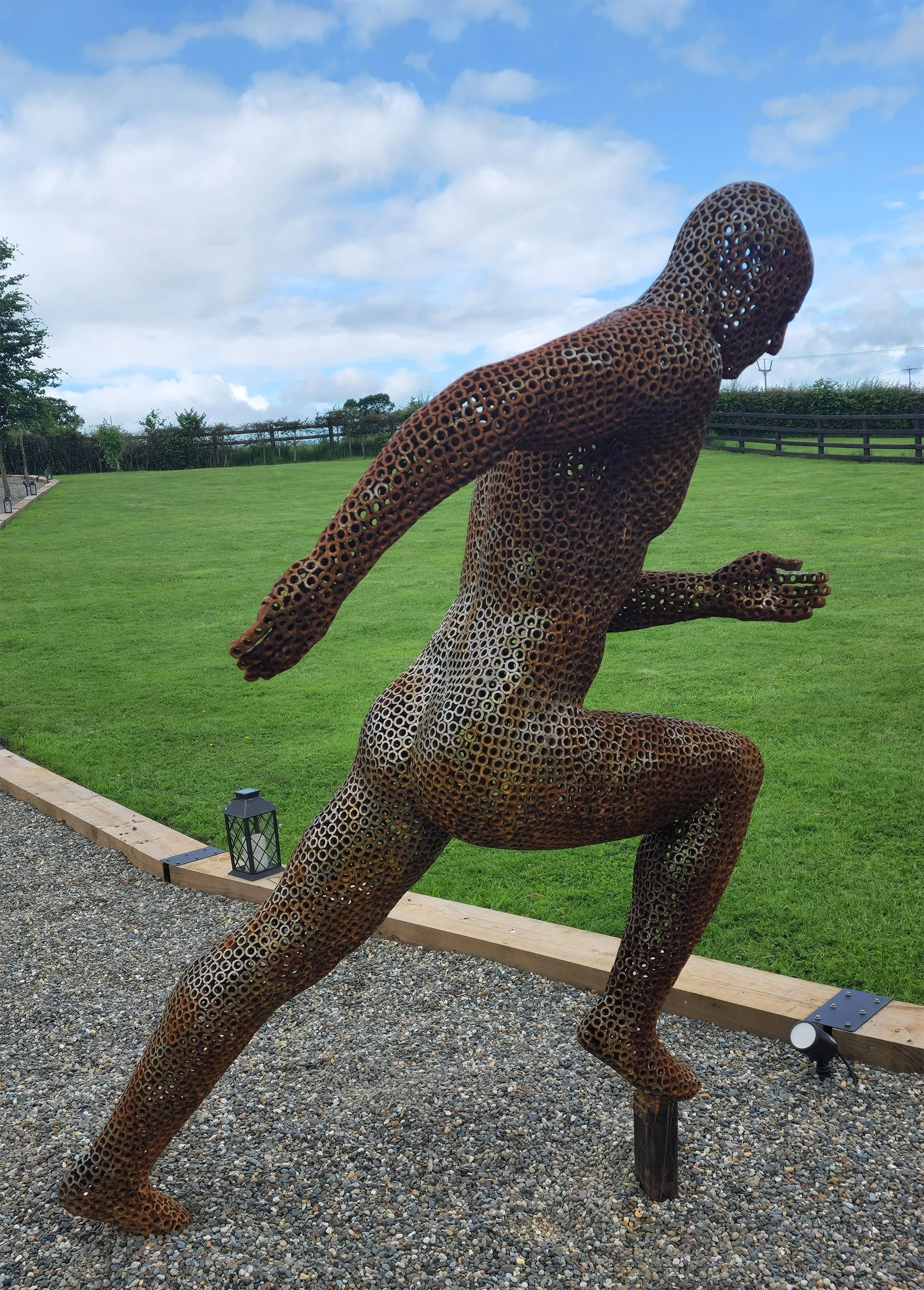A metal sculpture of a running man, designed by Shaun Gagg in a dynamic pose with one foot lifted off the ground. The figure is made of nuts, creating a semi-transparent, honeycomb-like structure.
