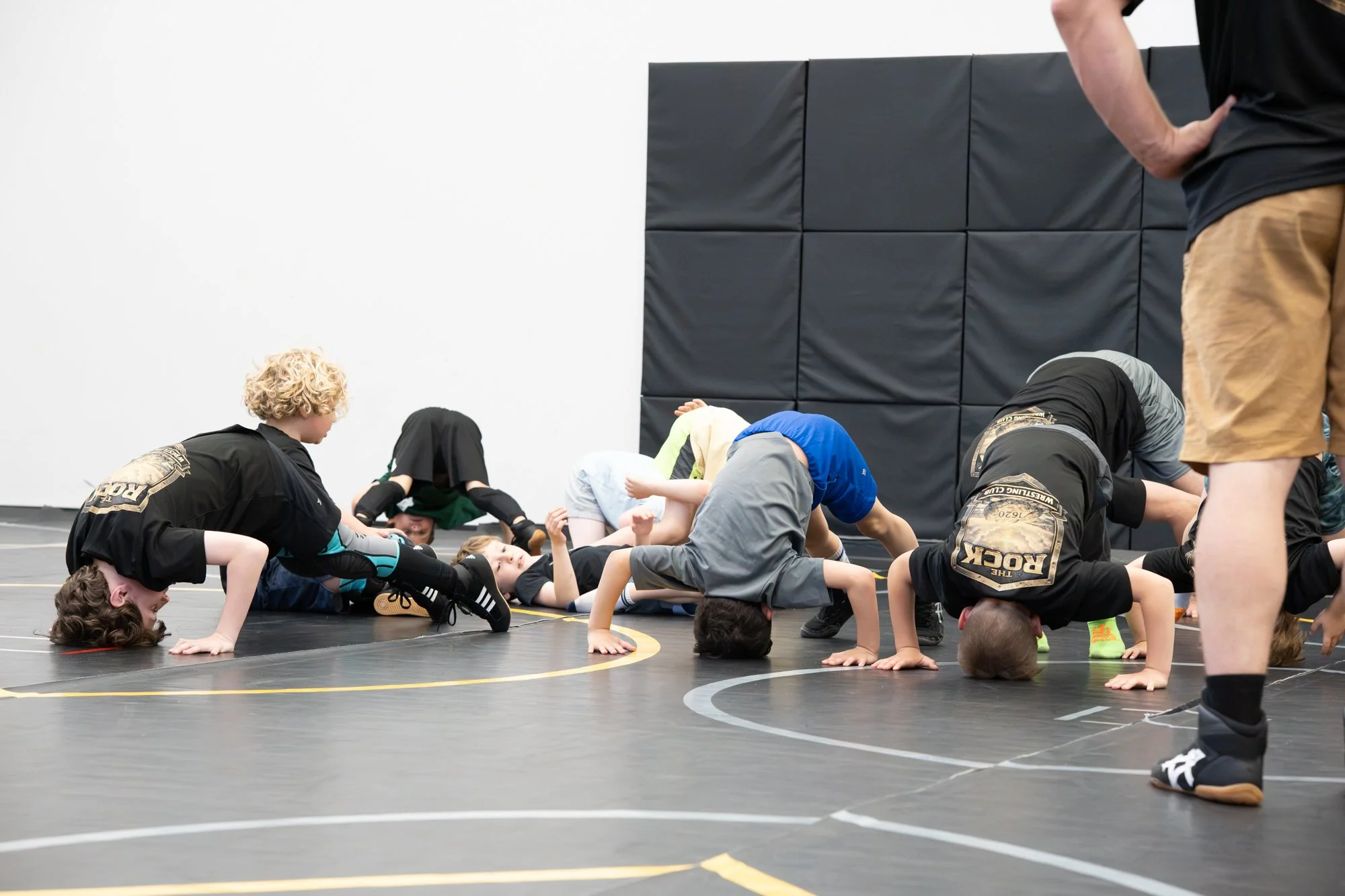 Children practicing wrestling techniques on a black wrestling mat in a gymnasium with an adult supervising.
