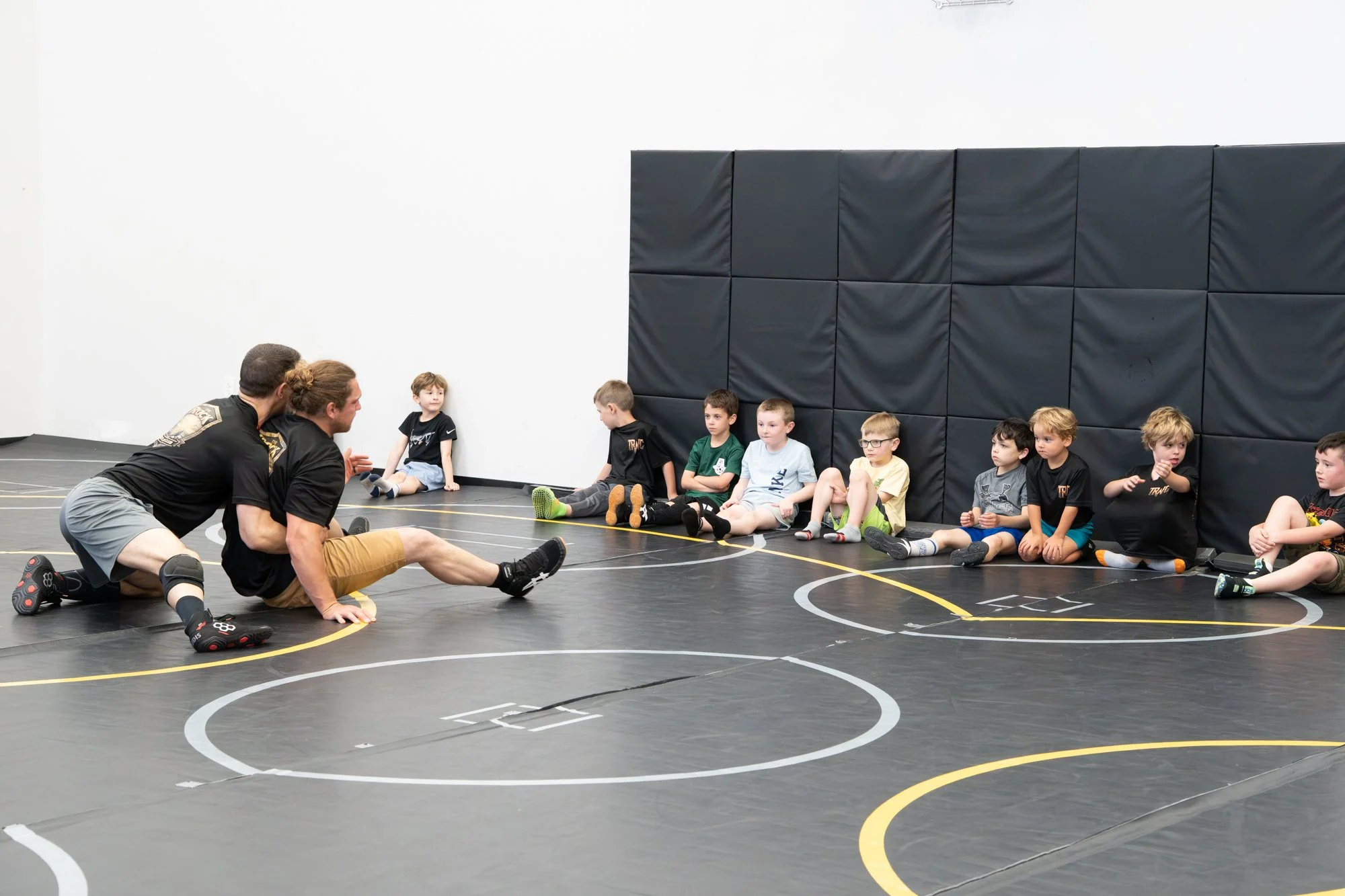 Two wrestling coaches kneeling on a black wrestling mat, speaking to a line of young boys seated against a padded wall during a wrestling practice or class inside a gym.