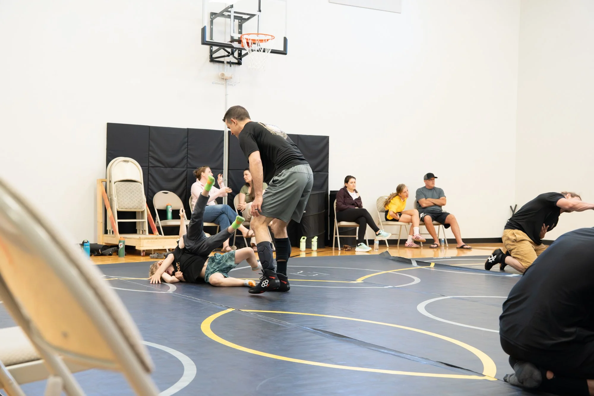 Children and adults participating in a wrestling practice or class in a gymnasium with chairs and spectators in the background.