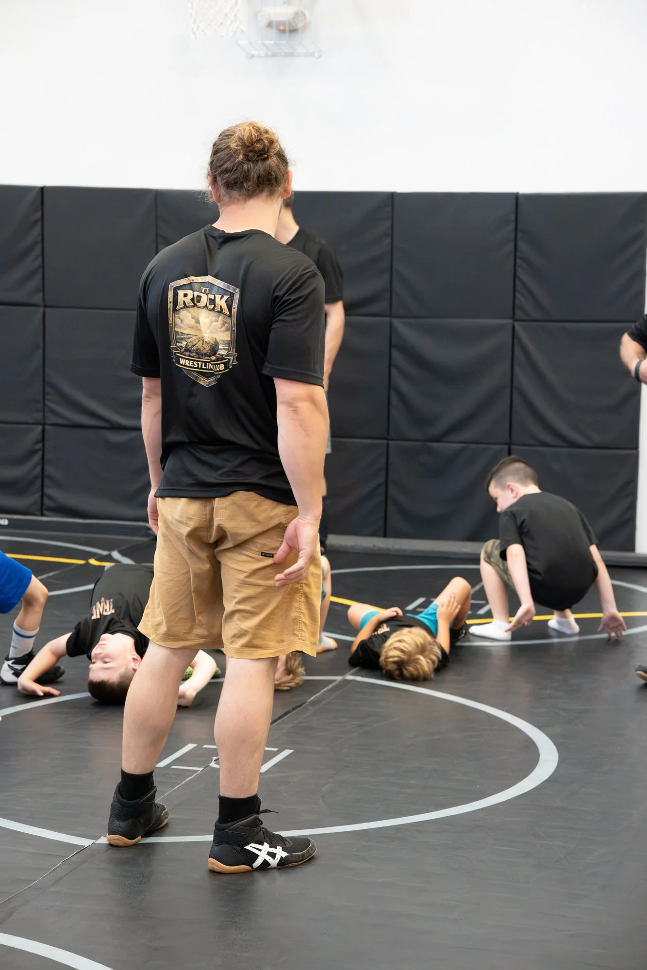 Children practicing wrestling moves on a wrestling mat at an indoor gym, overseen by passersby.