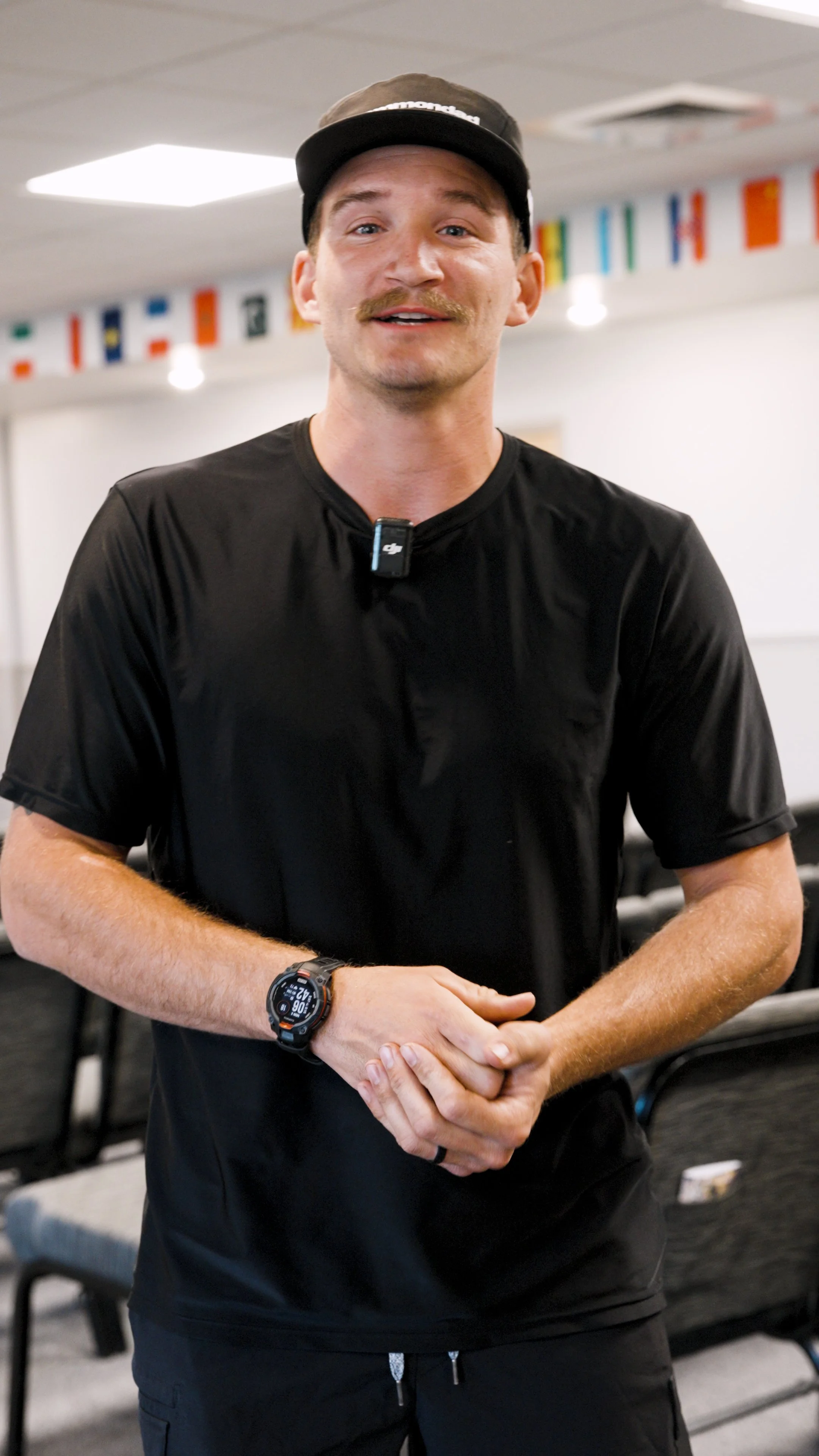 A young man with a mustache is smiling at the camera, wearing a black cap, black athletic shirt, and a wristwatch. He is standing with his hands clasped together in an indoor setting with flags hanging in the background.