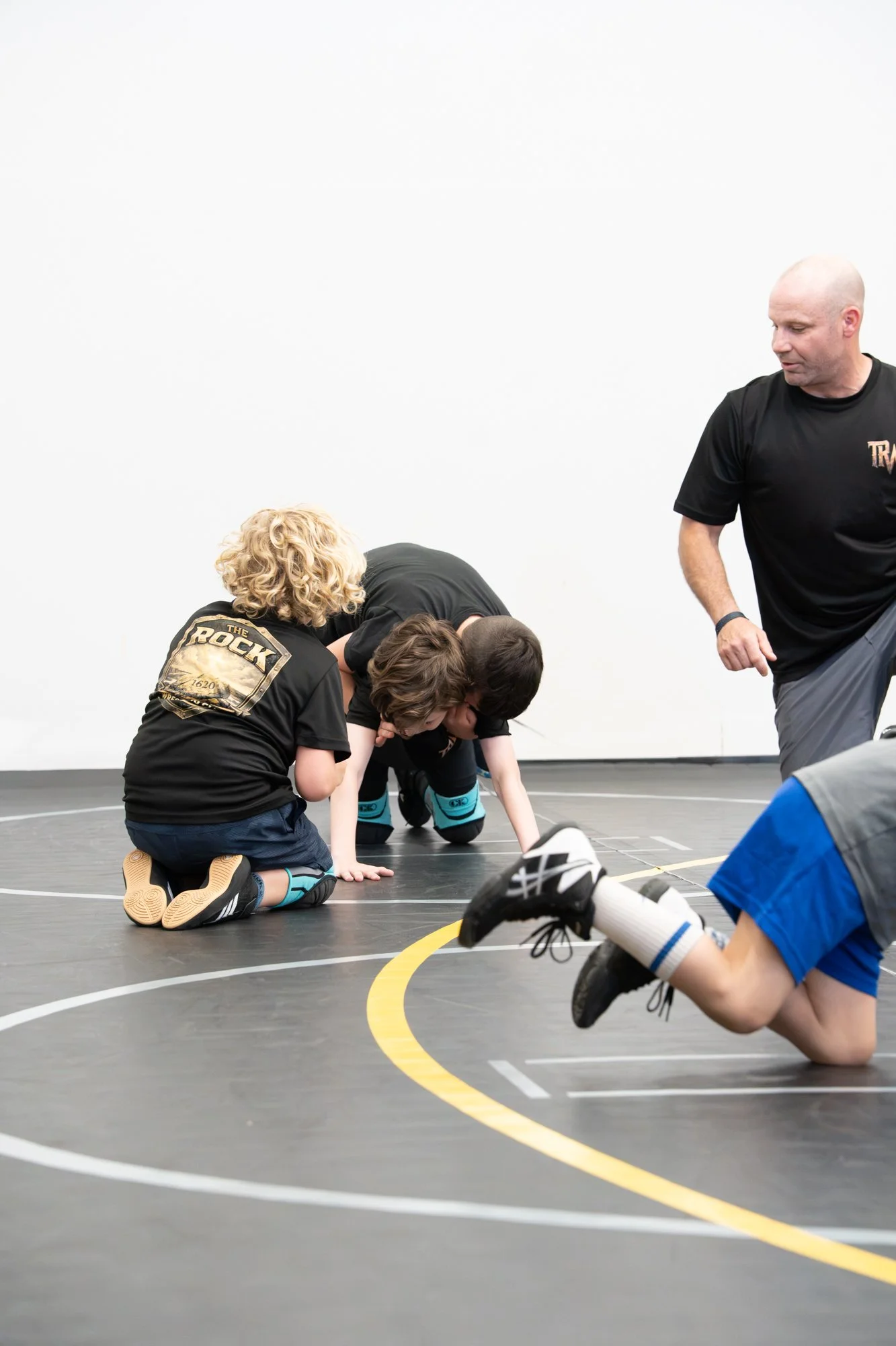 Young boys in black jerseys kneeling and crawling on a wrestling mat during practice with an adult coach standing nearby.