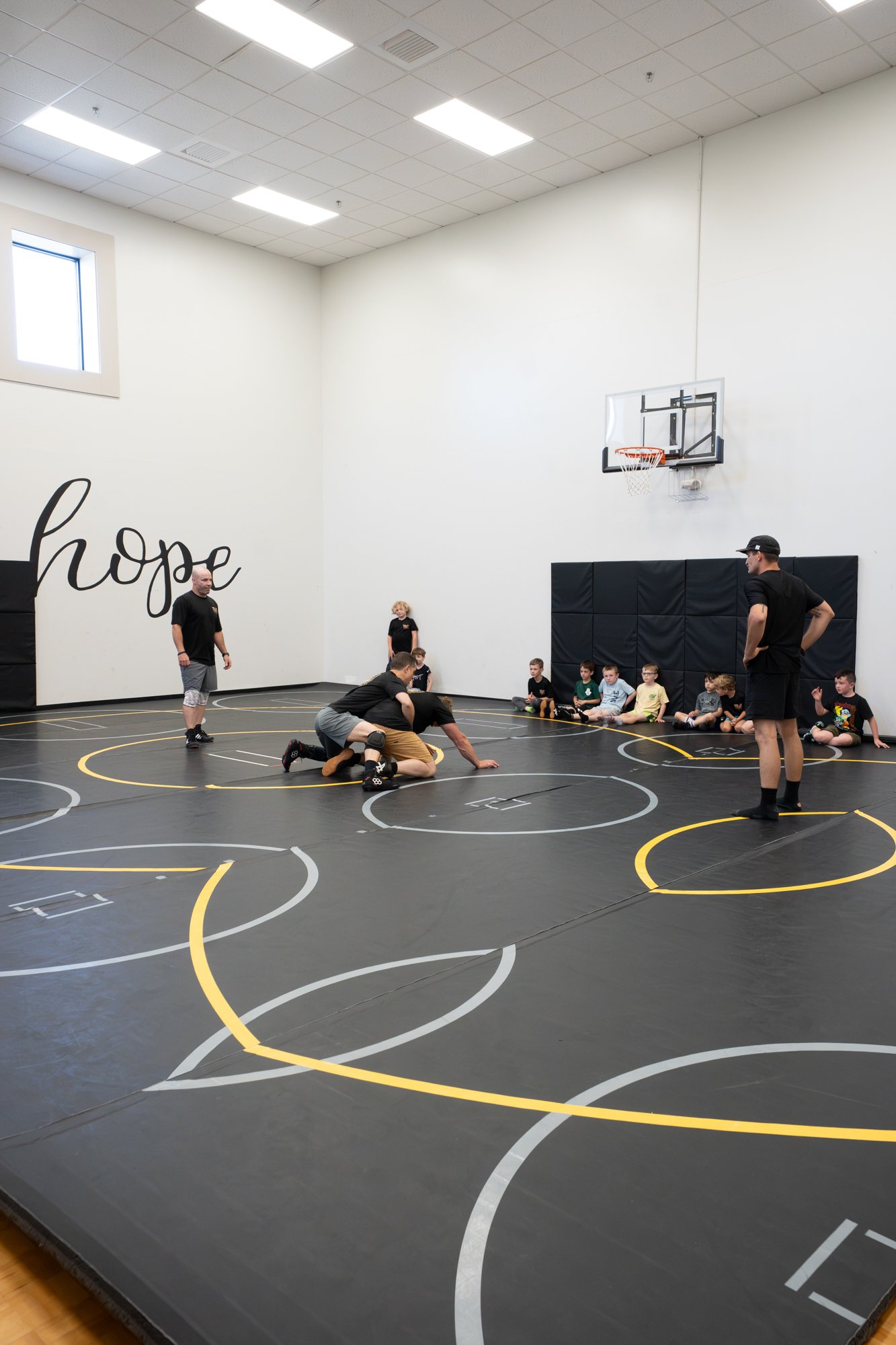 A gymnasium with a black wrestling mat marked with yellow and white lines. Several kids and adults are participating in a wrestling practice or class, with some kids sitting against the wall and others engaged in wrestling moves on the mat. The wall 