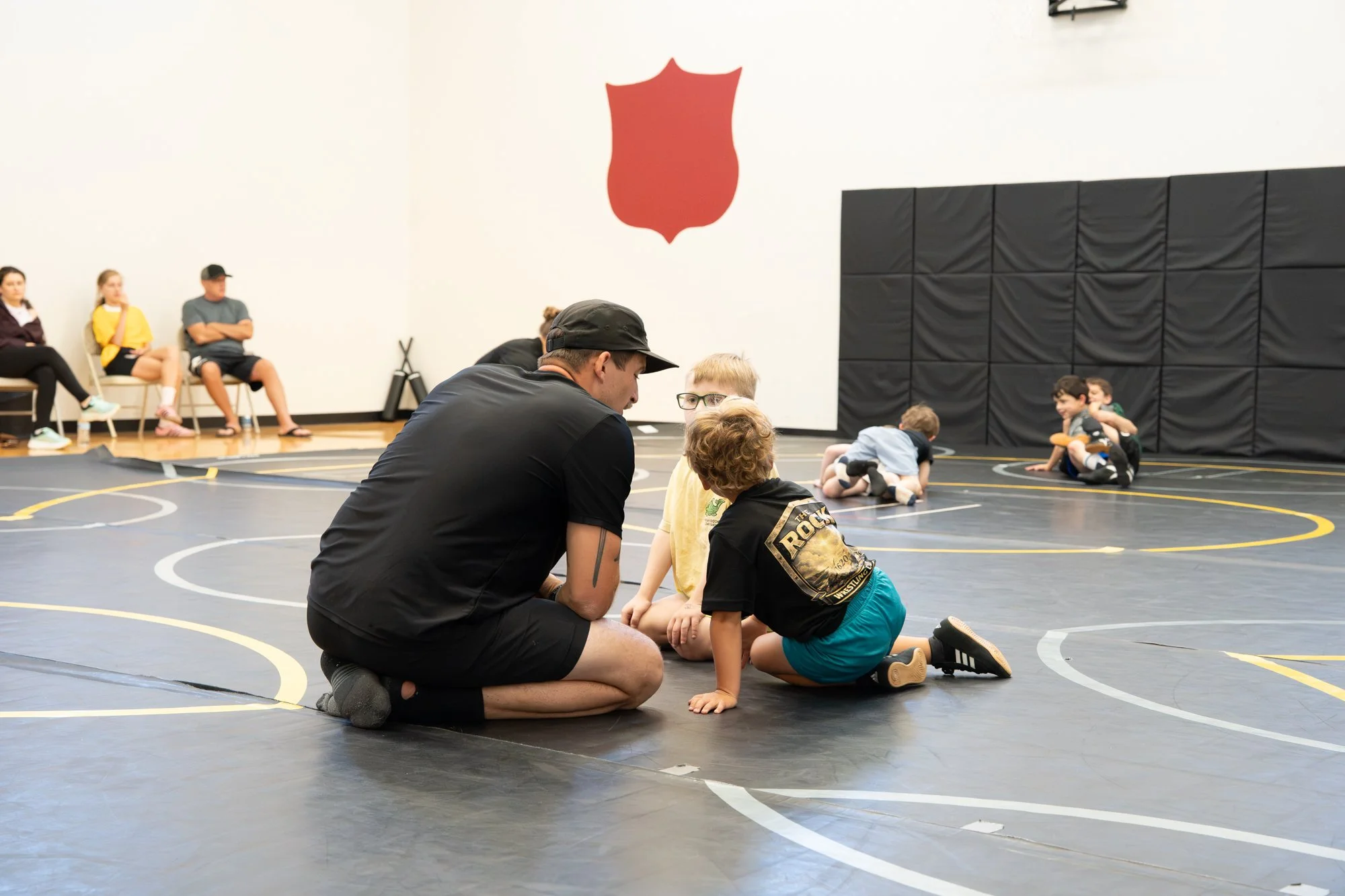 A wrestling event taking place on a black wrestling mat inside a gymnasium. A man with a black hat and black shirt is kneeling and talking to a young boy wearing glasses, a black t-shirt, and teal shorts. Several other children are seated on the floo