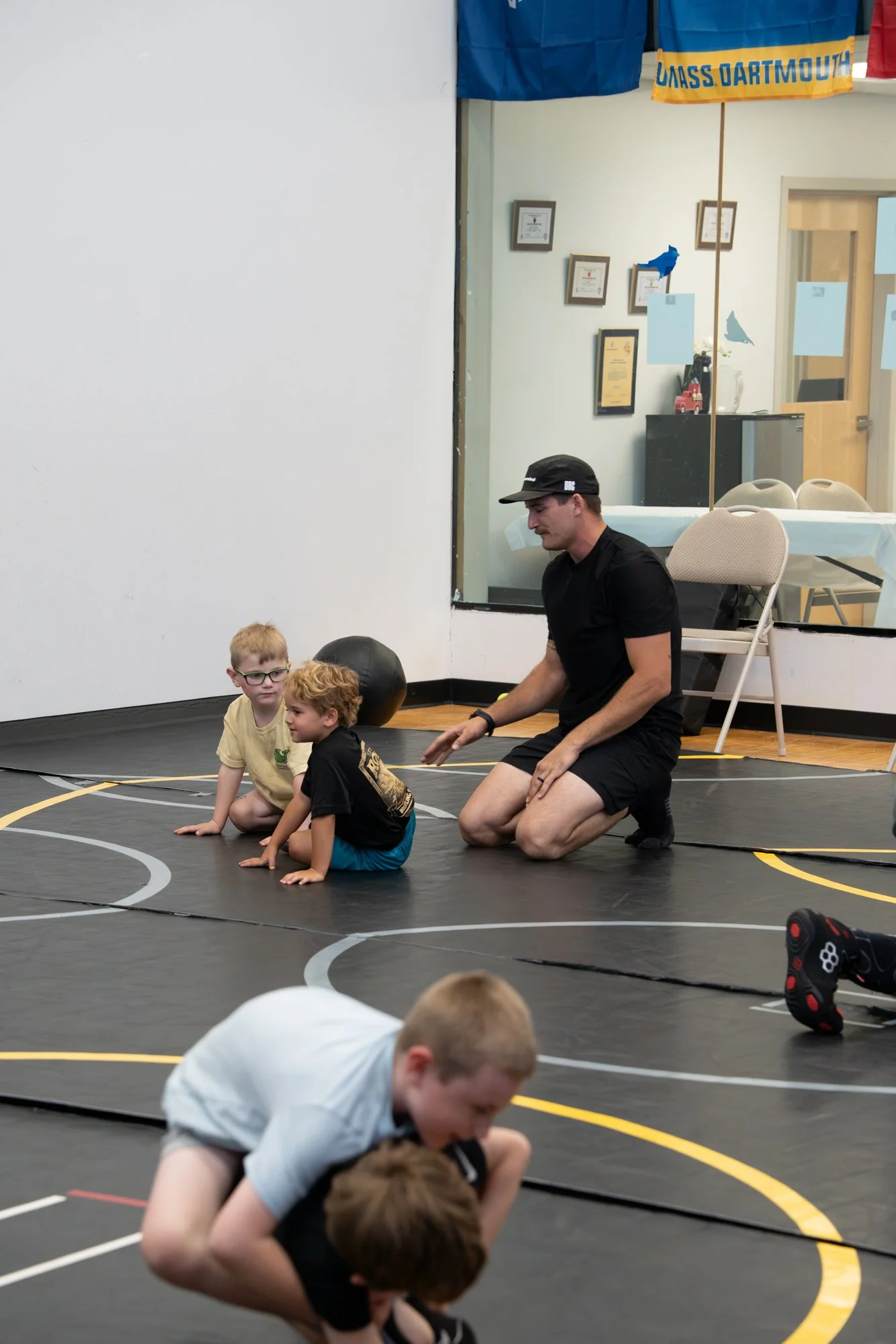 A coach kneeling on the mats instructs young children in a wrestling class, with some children practicing or resting on the mat, in an indoor gym with wall certificates and flags.