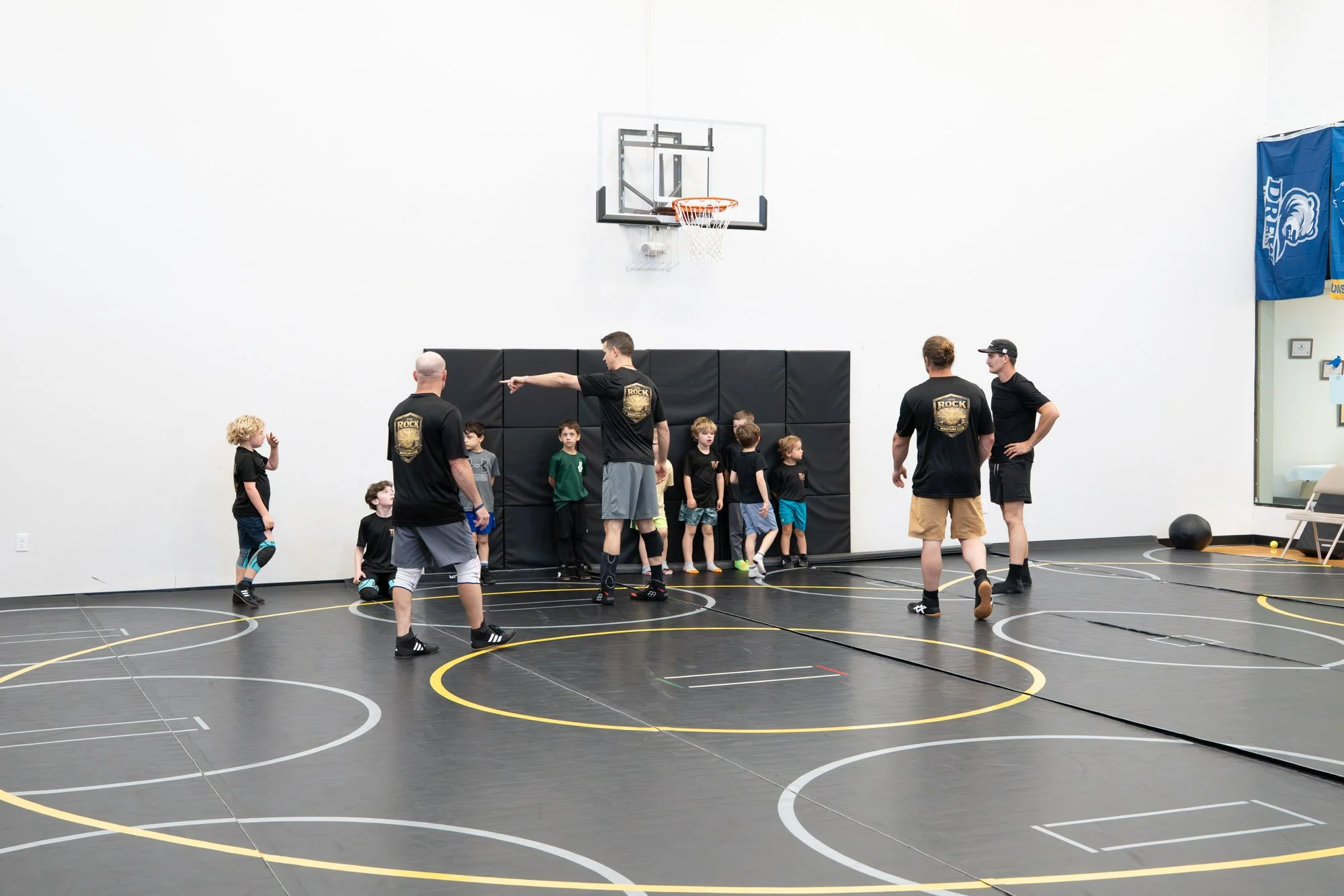 A group of children and coaches in a gymnasium with a basketball hoop. The children are standing in a line while the coaches give instructions. The gym has a black padded wall, a blue gym banner, and a digital clock on the wall.